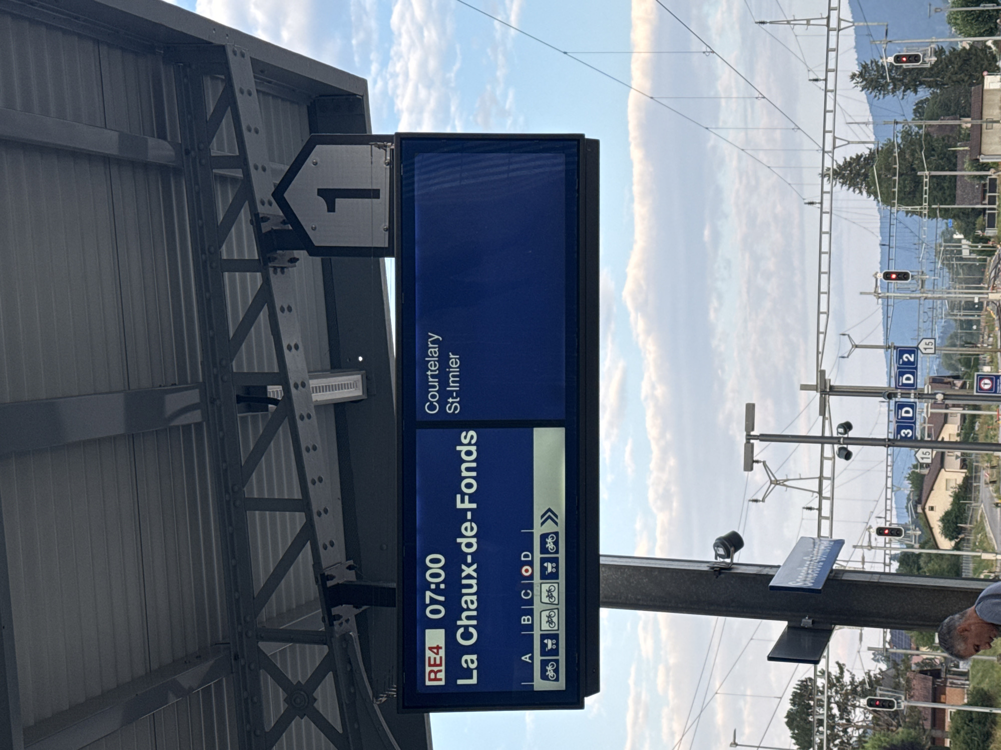 The image shows a train station platform in Sonceboz-Sombeval, Switzerland. A digital display board indicates that train RE4, departing at 07:00, is bound for La Chaux-de-Fonds and will stop at Courtelary and St-Imier. Below the destination information, symbols indicate available spaces for bicycles and wheelchairs on different train sections labeled A, B, C, and D. The platform roof is supported by a metal structure. In the background, railway tracks, overhead electrical lines, traffic lights, and buildings can be seen. A man is partially visible on the left side of the image. The sky is cloudy.