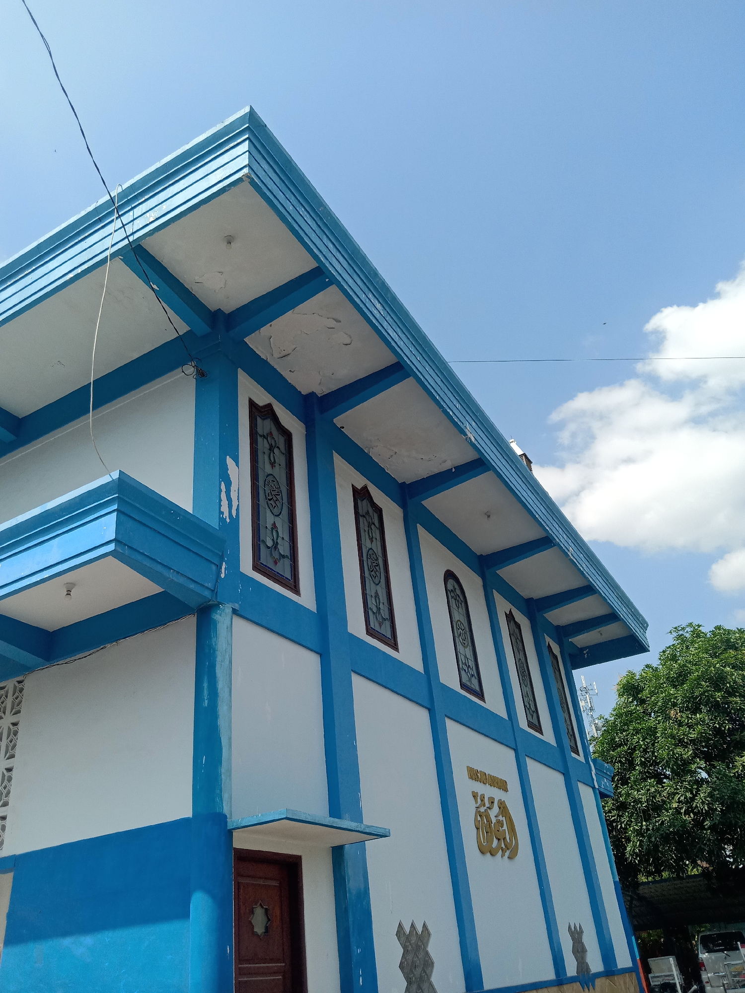 An upward-angle exterior shot captures the side of a white and blue building, possibly a mosque, in Sugihwaras, Indonesia. The building has a blue, stepped roof overhang supported by white columns. Several tall, arched windows with decorative stained glass are visible along the upper portion of the building's white facade, bordered by blue trim. Below these windows, on the white wall, is gold Arabic calligraphy, and further down, a dark wooden door is partially visible. The sky above is bright blue with scattered white clouds. To the right of the building, a large, leafy green tree stands, and some vehicles are parked under a dark awning in the background. The scene is shot from a low angle, emphasizing the architecture against the sky.