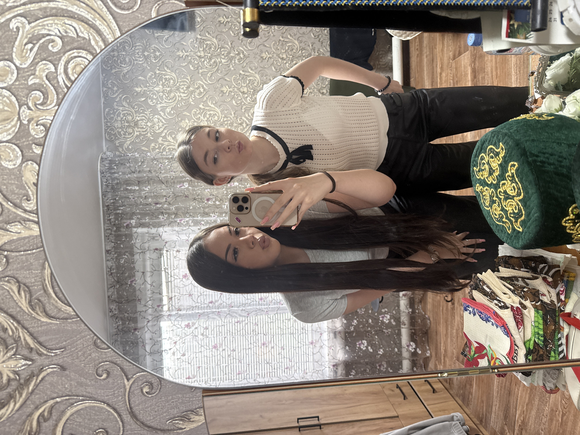 Two young women are taking a selfie in a mirror. One is holding a phone, her long dark hair hanging down. The other woman is standing behind her, wearing a white top with black trim. A green embroidered skullcap, various scarves, and a white shoe are visible next to them. The background includes wallpaper with a floral pattern and a window with patterned curtains. The location is Коноваловка, Russia.