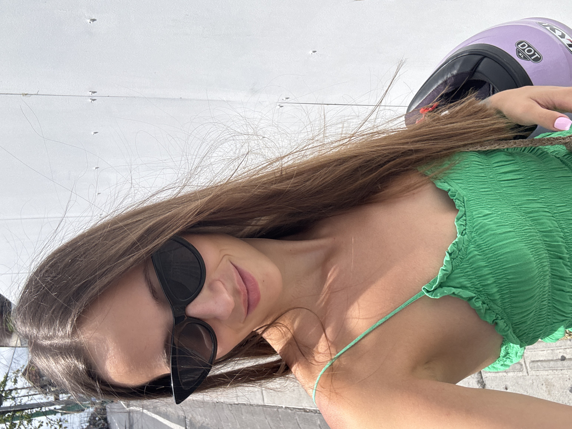 A woman wearing a green top and black sunglasses is holding a purple helmet. The location appears to be in the city of North Kuta, Indonesia.