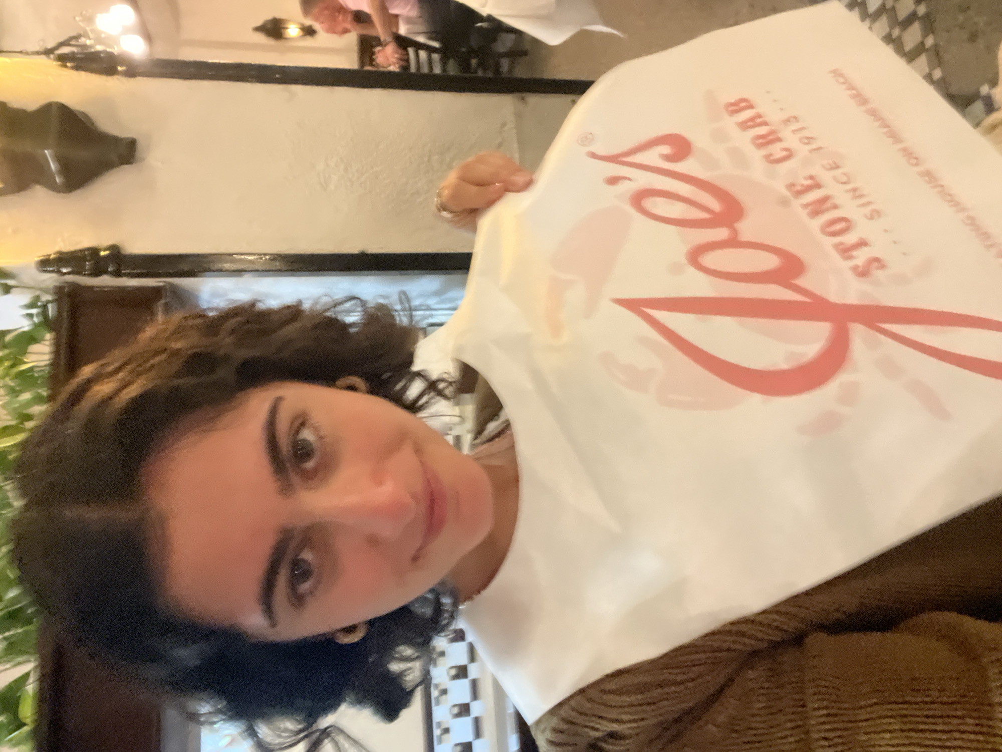 A woman is seated at a table indoors and holding up a bib from "Joe's Stone Crab" which is located in Miami Beach, Florida, United States. The bib is white with red lettering. She is wearing a brown sweater. A person is seated at a table in the background. A light fixture and molding are visible on the wall. A plant is visible to the left of the woman.