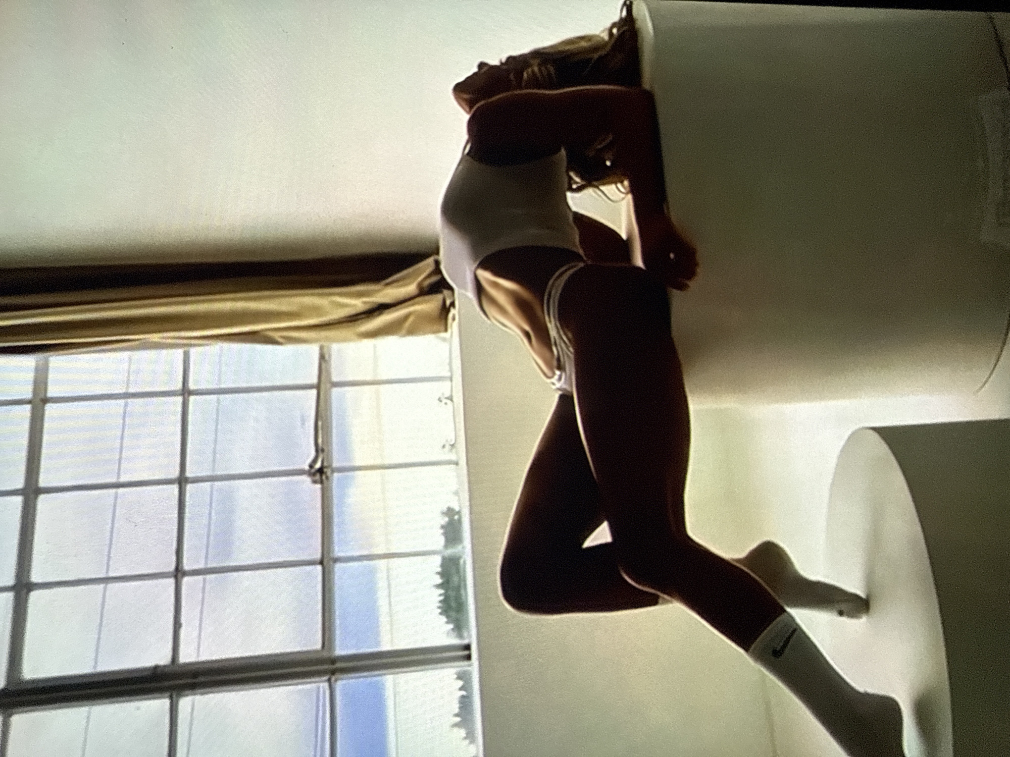 This image depicts a studio setting, identified as being in Varna, Bulgaria. A lean individual, appearing to be a female, is positioned in an arched back pose across two distinct white cylindrical platforms. The person wears a white tank top, white athletic-style underwear with thin horizontal stripes, and white socks, one displaying a dark Nike logo on the ankle. Her head is tilted far back, allowing long, light brown hair to flow downwards. Her elbows rest on the lower, wider platform, supporting her upper body, while her legs are extended onto the taller, narrower platform, with one foot pointed and the other slightly bent. In the background, a large industrial-style window, segmented by dark frames, reveals a bright sky and distant green foliage. A gold or tan curtain is visible to the right of the window. The overall environment is brightly lit and minimalist.