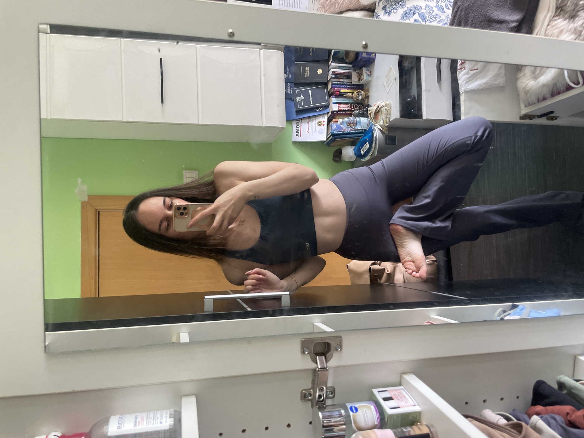A woman is reflected in a mirror, taking a selfie with a smartphone. She is wearing a dark sports bra and gray leggings. The reflection shows a room interior, including a green wall, a light wood door, white cabinets, and items on a shelf above the mirror, such as books and toiletries. Based on the presence of Bulgarian language materials on the shelf, the location is possibly in Sofia, Bulgaria.