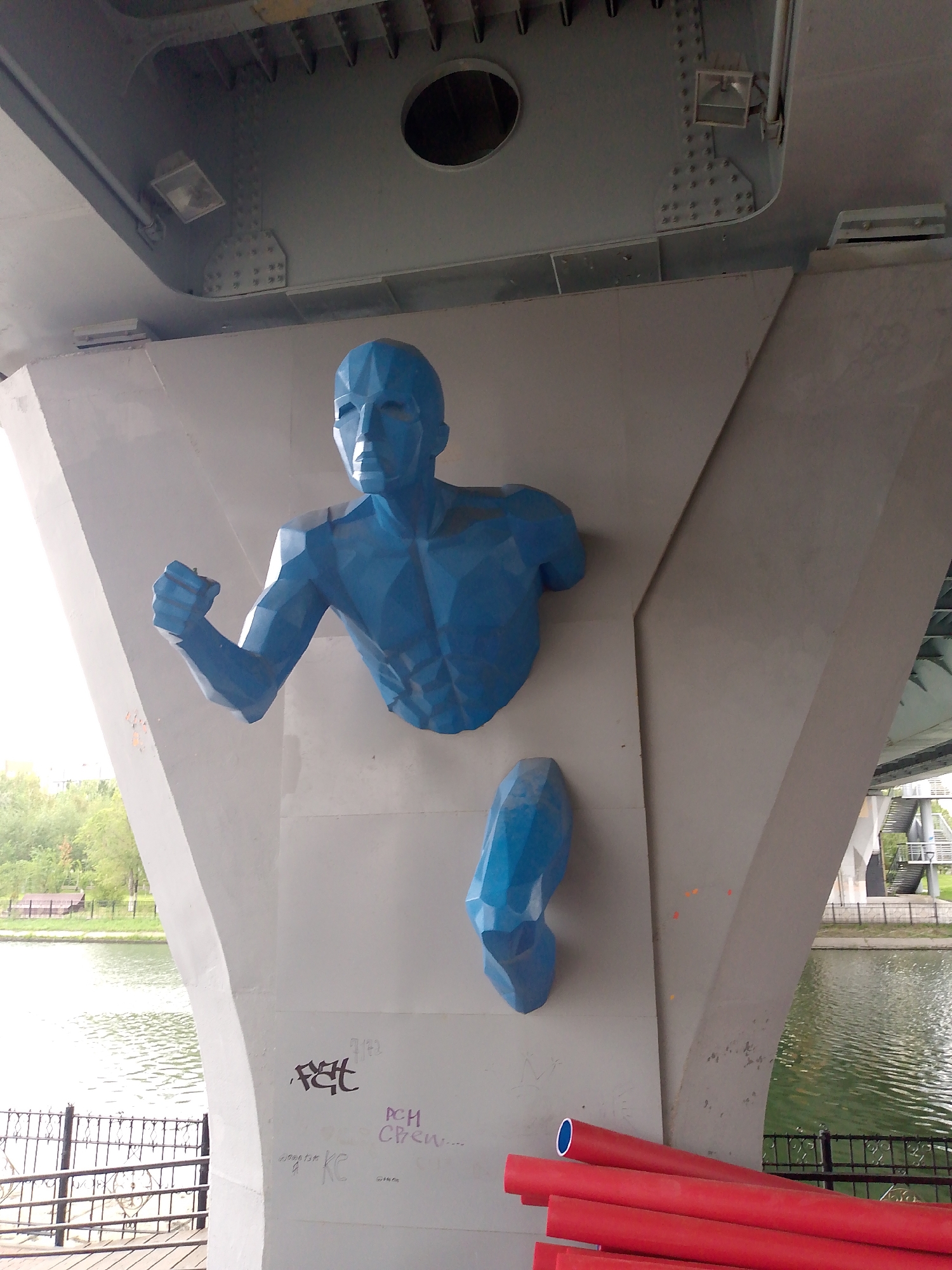 A blue, polygonal sculpture of a human torso and leg is mounted on a large concrete support structure under a bridge. The sculpture depicts a figure in a running pose, with its left arm bent and fist clenched, and its right leg detached and positioned below the torso. The sculpture is made of geometric planes and has a matte finish. The concrete support is a light gray color and has some graffiti scribbled on its surface. Below the sculpture, several red, cylindrical objects are piled on top of each other. In the background, a body of water is visible, with trees and a fence along its edge. The scene appears to be outdoors in an urban environment. The sculpture is known as "Runner" and is located on the Astana embankment in Kazakhstan.