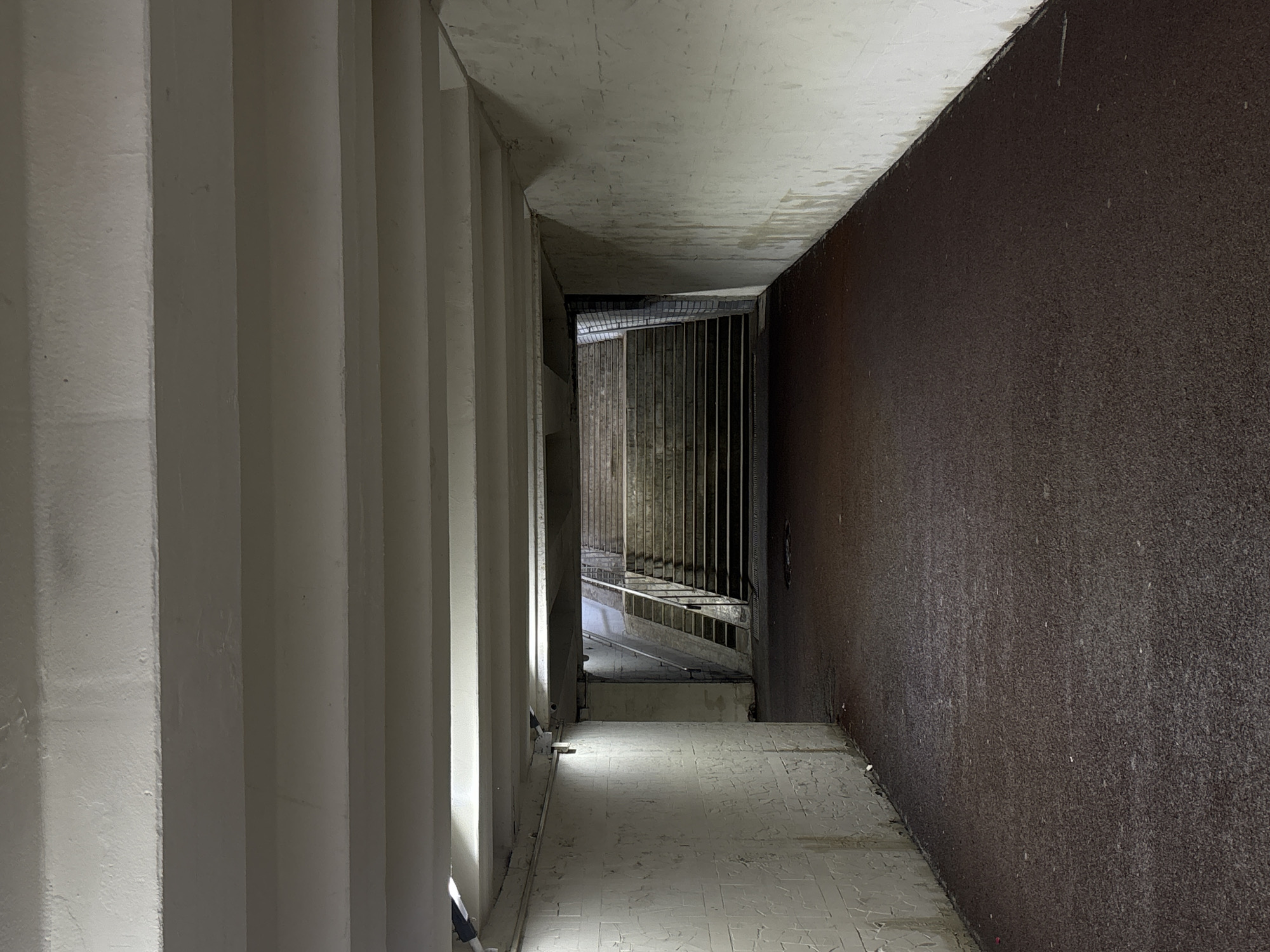 The image depicts a narrow, dimly lit corridor, likely a pedestrian underpass, in Perm, Russia. The left wall consists of a series of vertical, white concrete pillars. The right wall is a rough, dark brown textured surface. The floor is light gray concrete with a tiled pattern. Further down the corridor, a metal grate fence with handrails is visible, beyond which the corridor continues. The ceiling is exposed concrete. There is no indication of any people present in the image.