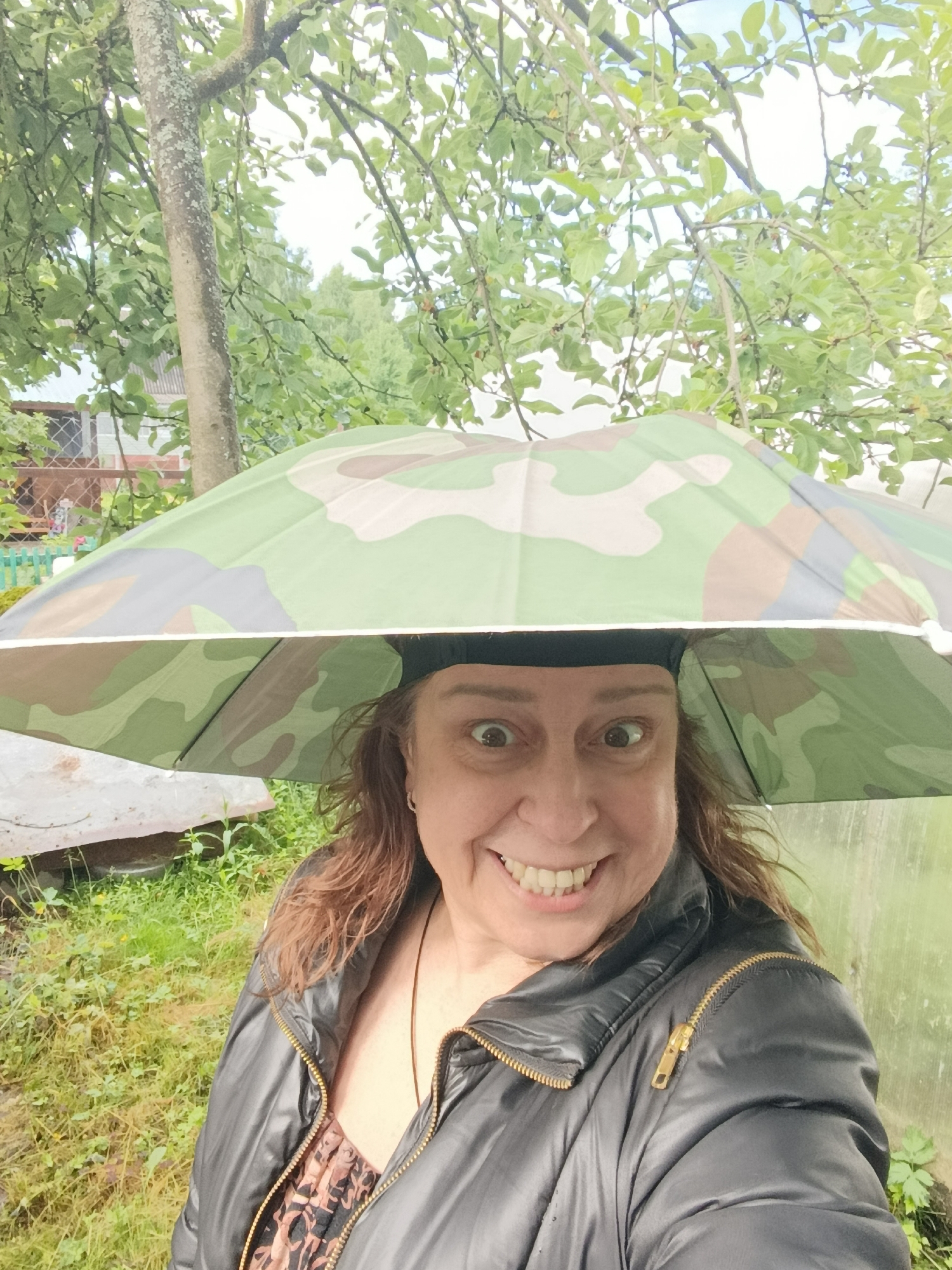 A woman stands outdoors, holding a camouflage umbrella over her head. She is wearing a black puffer jacket with a gold zipper and a patterned top underneath. Her expression is wide-eyed and she is smiling. The background shows trees with green leaves, tall grass, and a fence. The location is likely a residential area in Verëya, Russia, given the presence of trees, gardens, and a fence, common features in such settings.