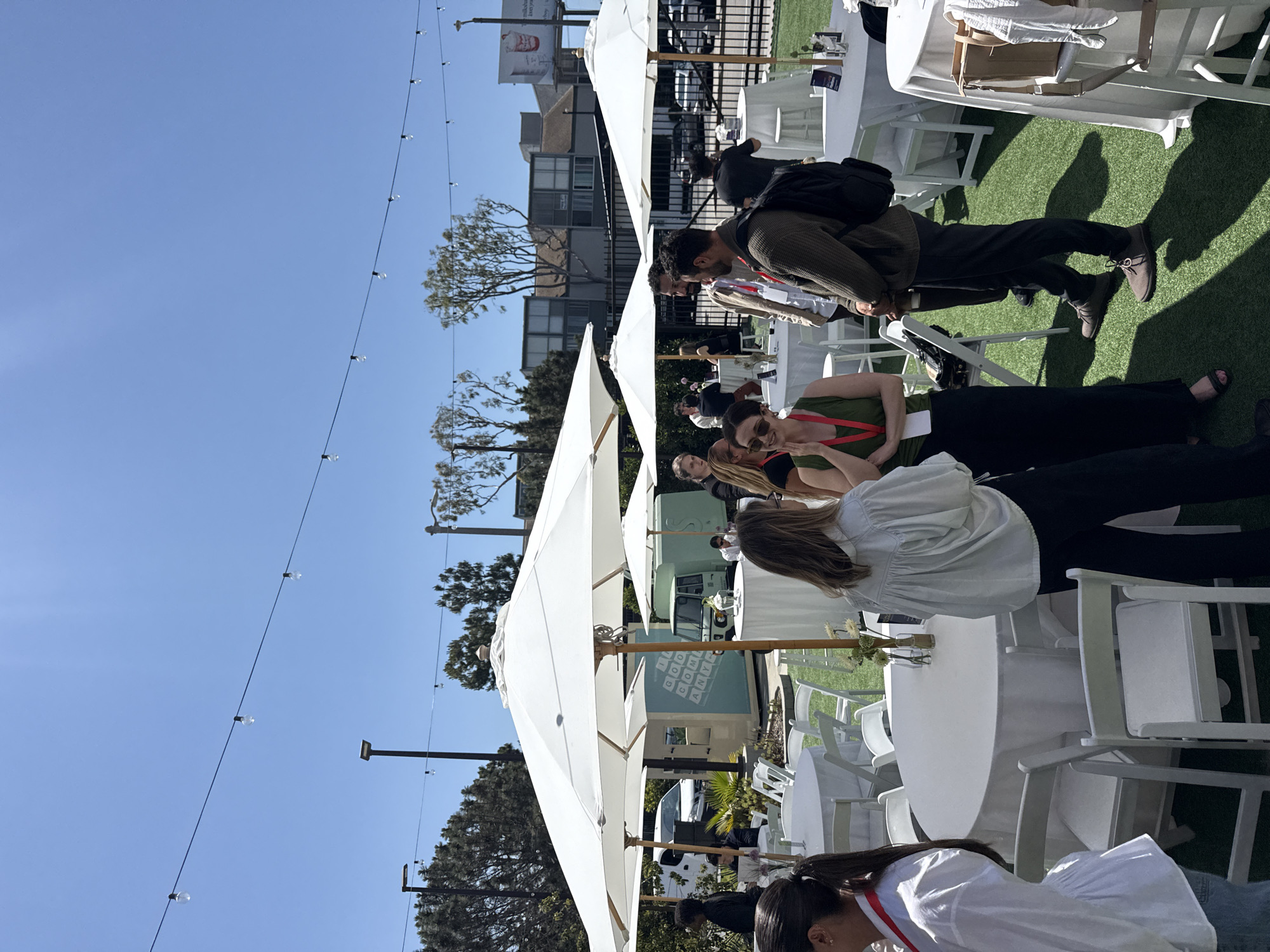 The image shows an outdoor event under a clear blue sky with string lights overhead. Several people are present, sitting at round tables with white tablecloths and chairs under large white umbrellas. The event appears to be taking place on artificial turf. Behind the tables, there are buildings, trees, and other urban elements, including a sign referencing Los Angeles. It seems to be a gathering or conference of some kind. The location is confirmed to be in the city of Los Angeles, United States of America.
