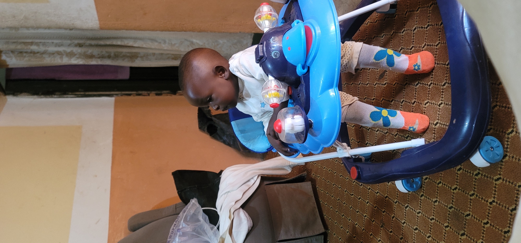 A young child is in a blue and white baby walker with wheels on a brown patterned carpet in an indoor location. The child wears a white shirt and floral-patterned socks with orange toes and heels. In the background, partially visible, is a wall with alternating light and dark orange stripes, a curtain, and some dark-colored furniture. This scene is located in Barakin Akawo, Nigeria.