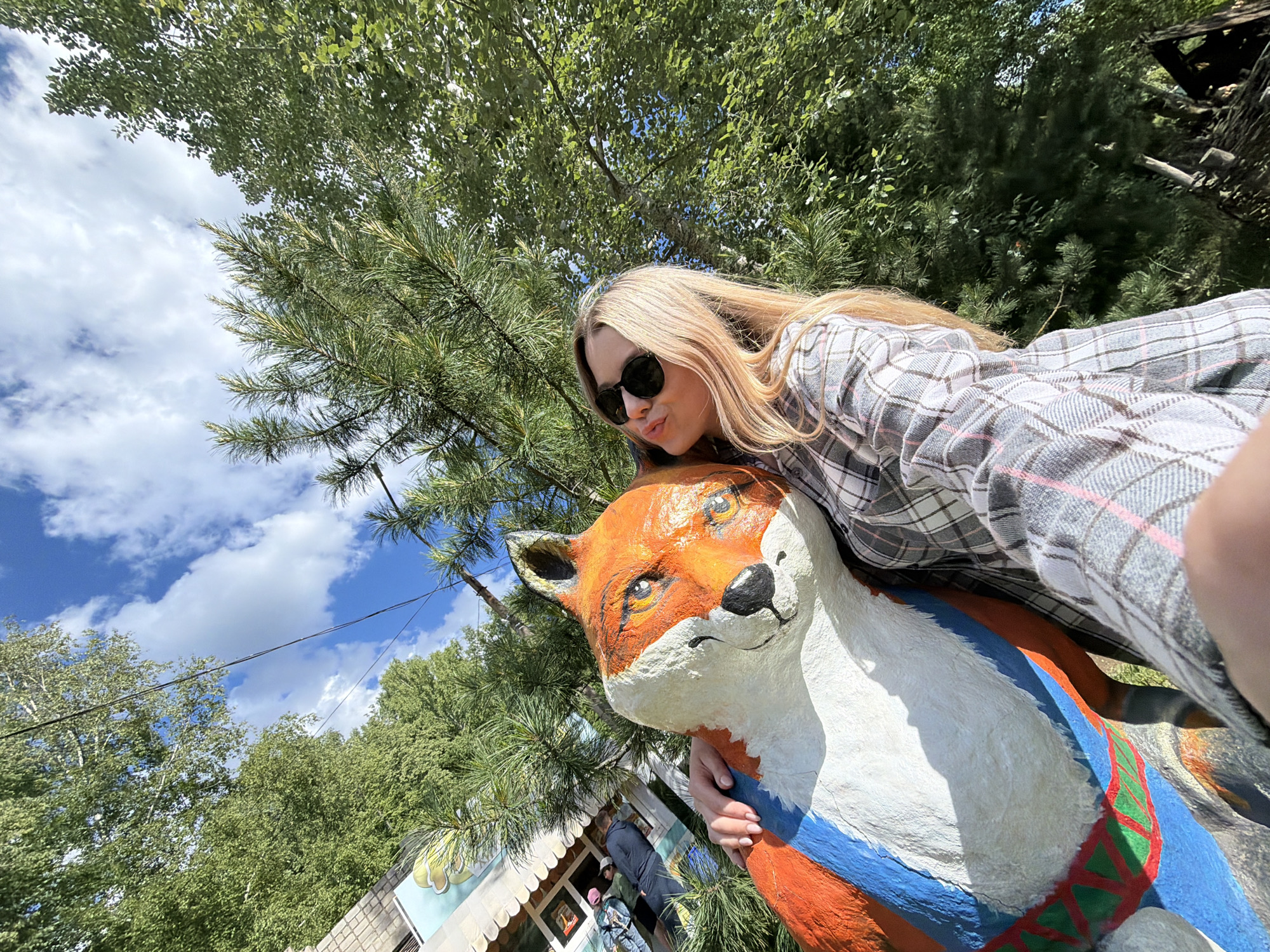 A low-angle, wide shot captures a woman posing with a painted fox statue in Chaikovsky, Russia. The woman, fair-skinned with long blonde hair and wearing sunglasses and a gray plaid shirt, leans toward the fox with puckered lips as if to kiss it. She is partially obscuring the fox. The fox statue is painted orange, white, and blue with a decorative element around its chest. Behind the woman and the statue, lush green trees fill the upper part of the frame. To the left, there's a glimpse of a building with a light-colored facade and what appears to be an awning. The sky is visible between the trees, featuring fluffy white clouds against a blue background. The overall perspective indicates the photo was taken from below, angling upwards toward the subjects. People are faintly visible inside the building next to the statue.