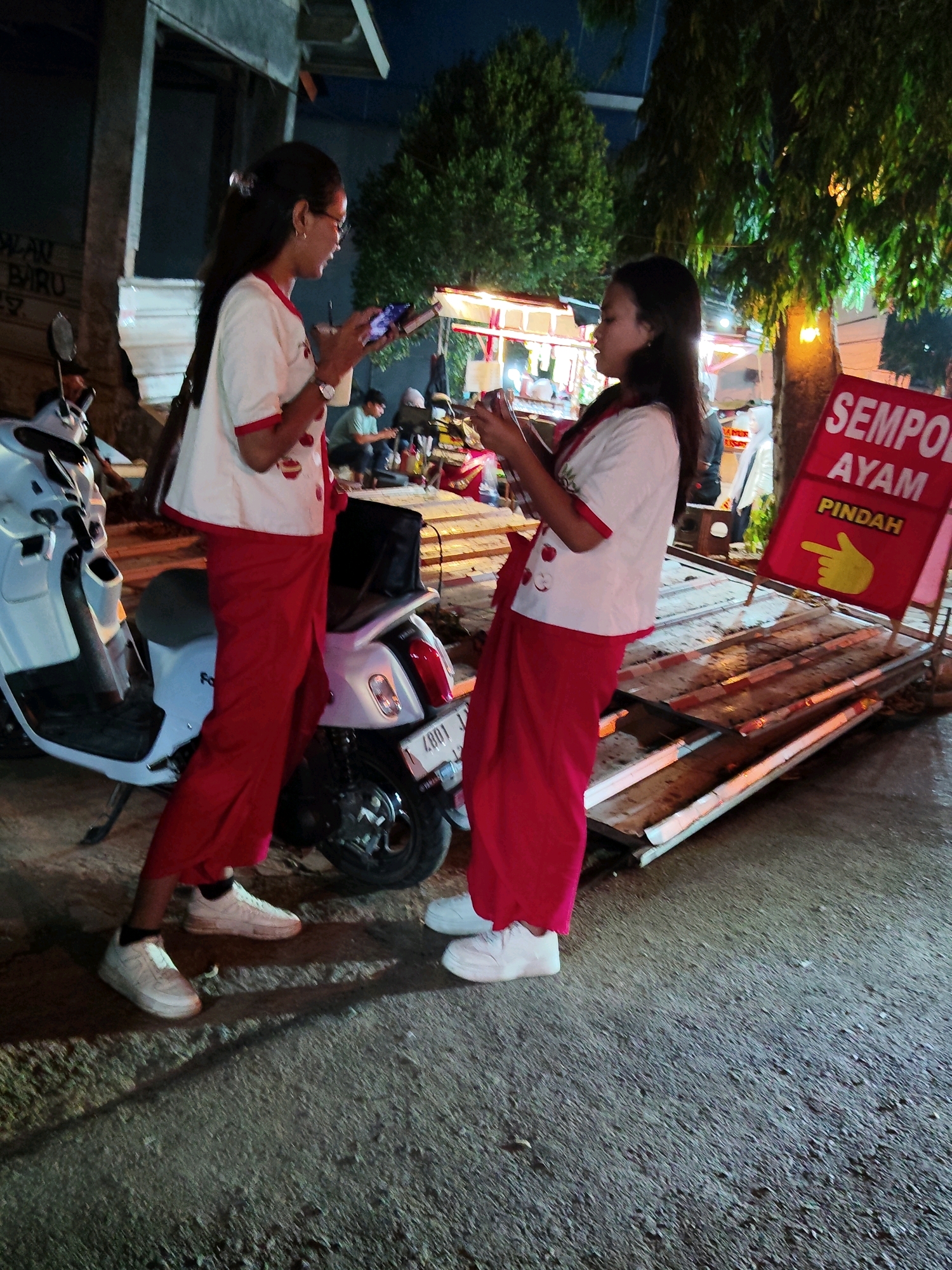 In Purwakarta, Indonesia, two young women dressed in matching red pants and white tops with red trim are standing next to a white scooter at night. The woman on the left, wearing glasses, is holding a phone and a drink. The woman on the right is also holding a phone. Behind them, a red sign with yellow lettering reads "SEMPO AYAM PINDAH," indicating a food vendor. A wooden platform is set up in front of the sign. The ground is a rough, dark asphalt surface. Trees with dense foliage are visible in the background.