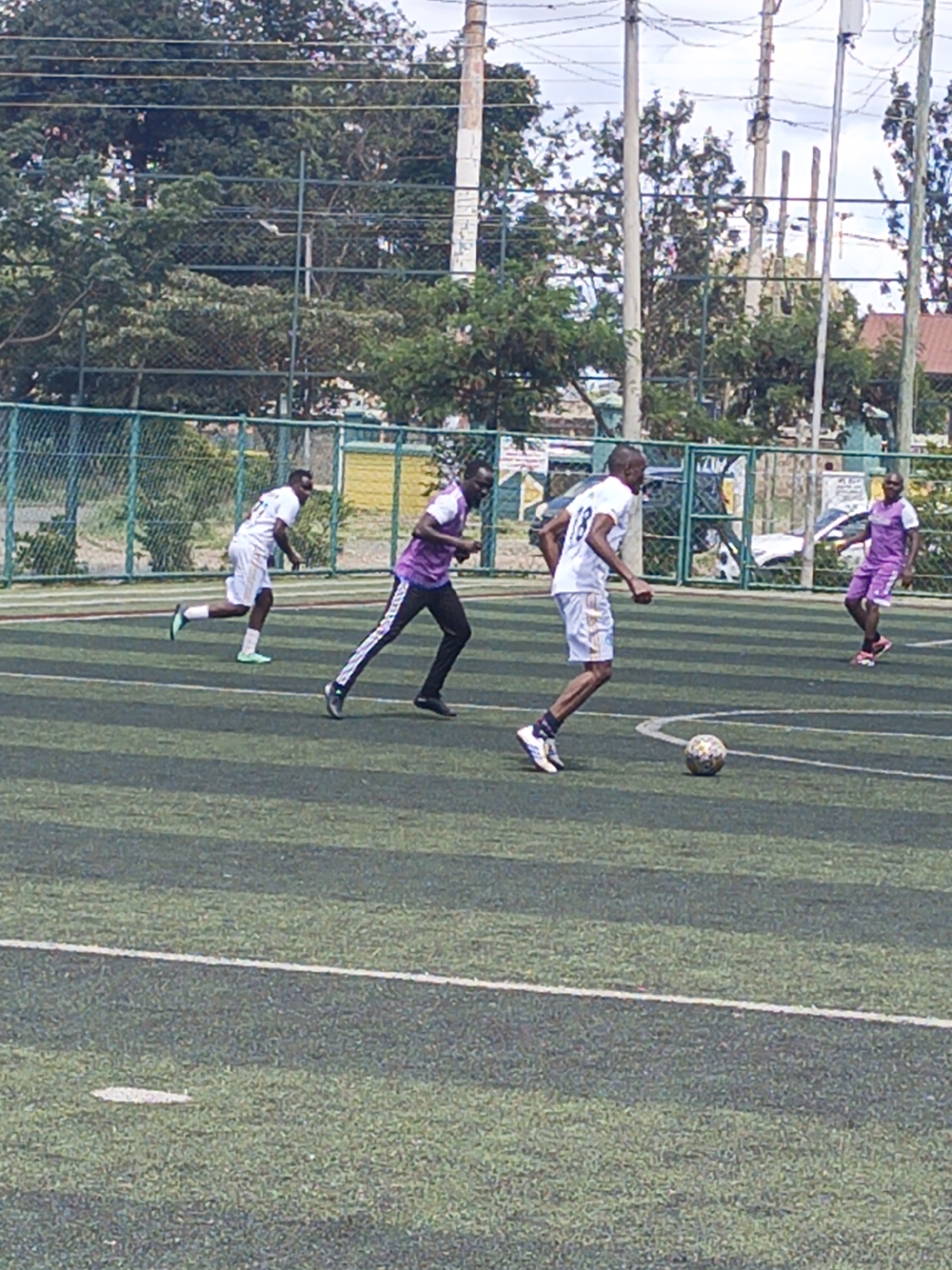 The image captures a soccer game in progress on an artificial turf field, likely in Nairobi, Kenya. Four men are visible on the field, engaged in the game. One man in a white jersey with the number "18" is actively dribbling a soccer ball. A man in a purple shirt is running to his left. Another player in white jersey, is running to his left and the fourth player is running to his right wearing purple jersey. A green fence runs along the edge of the field, and trees and utility poles are visible in the background.