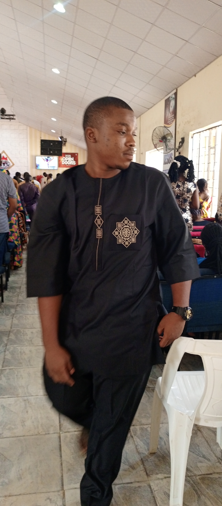 A man in a black embroidered outfit stands indoors in what appears to be a church. He is wearing a watch on his left wrist. In the background, other people are seated or standing. A white plastic chair is to his right. A ceiling fan and religious posters hang on the wall. The location is likely Keffi, Nigeria.