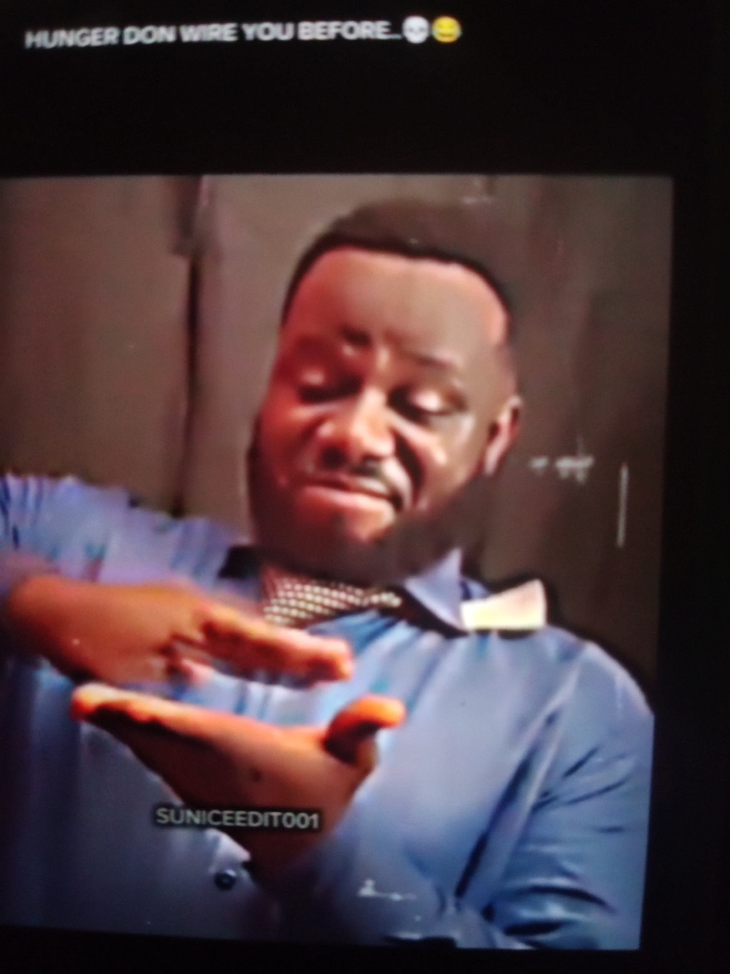 The image is a low-resolution, possibly screen-captured video still, showing a Black man in a blue collared shirt and a patterned bow tie. He has short, dark hair and a beard. His expression appears distressed or contemplative, with his eyes closed and his head tilted slightly downwards. His hands are held in front of him, with his right hand appearing to be gesturing towards his left palm, which is cupped. A watermark reading "SUNICEEDIT001" is visible on the man's shirt, near his hands. The background is a blurred, indeterminate, possibly indoor setting with a textured wall. The top of the frame displays white text that reads "HUNGER DON WIRE YOU BEFORE.. 💀😂".
There is no visible information within the image to suggest a specific location in Jalingo, Nigeria. The provided context about the city is not supported by the visual evidence.