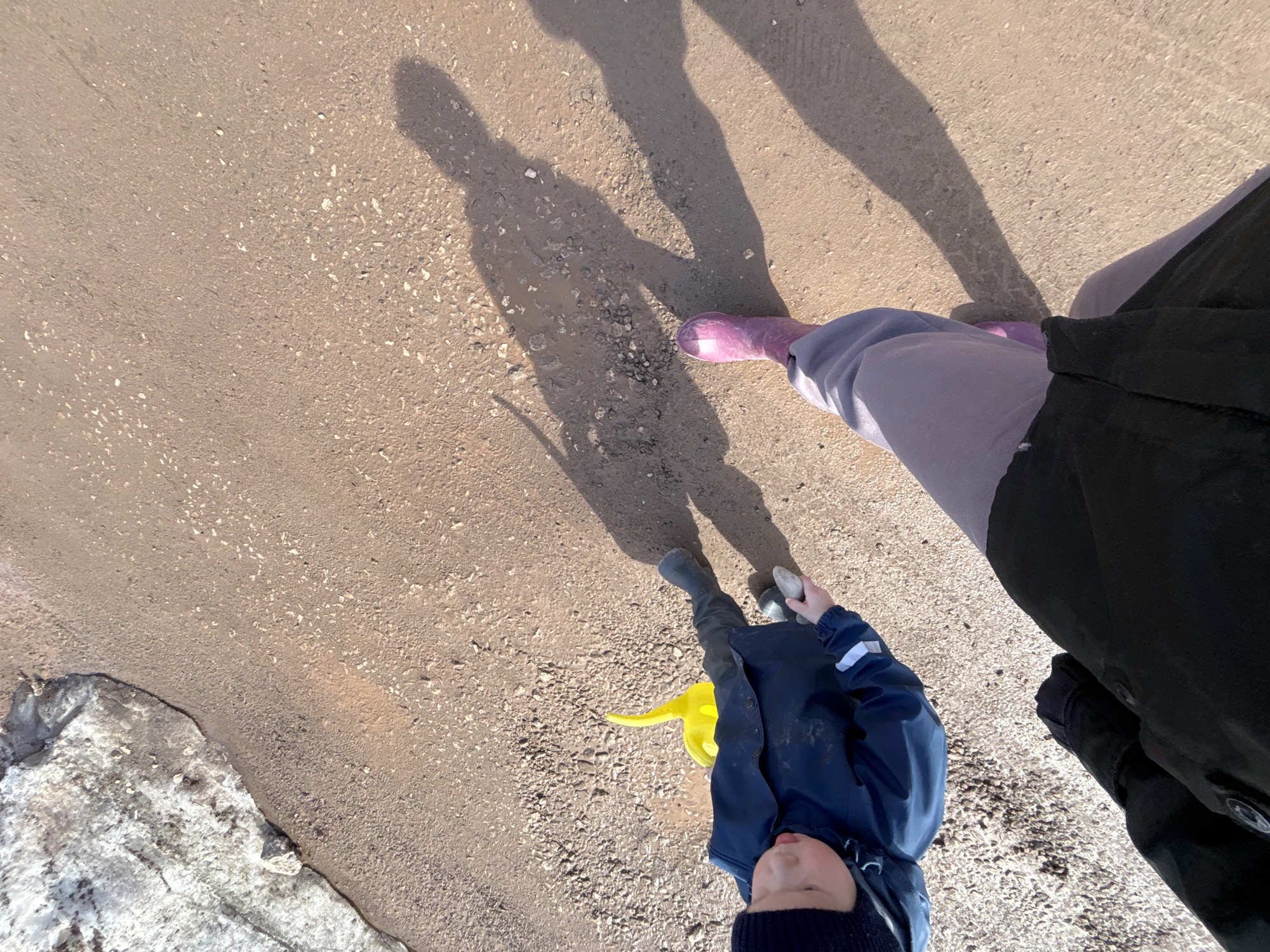 The image depicts a downward-angled view of a paved surface, likely a street or walkway, near Ферма, Russia, indicated by the presence of melting snow/ice along the edge of the pavement. Three individuals are partially visible, suggested by the shadows and partially visible bodies. A child in a blue jacket and dark blue knit cap is holding a yellow watering can. An adult wears purple rain boots and gray pants. Another adult wears black pants and a black coat. The individuals appear to be walking on the paved surface, as indicated by their shadows.