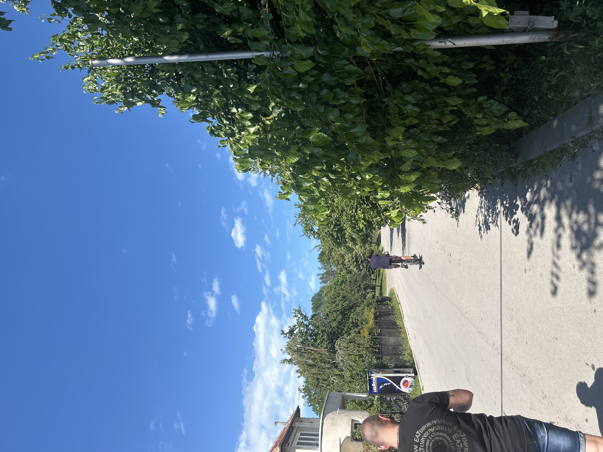 The image depicts a street scene in Varna, Bulgaria, under a clear blue sky with scattered white clouds. In the foreground, a person wearing a black t-shirt with a circular graphic design and denim shorts is walking away from the camera. Further down the asphalt road, another person is riding a bicycle, also moving away from the viewer. The road is lined with lush green trees and hedges, with a house visible on the left side of the street. A vending machine with a "Lavazza" logo is positioned near the house. Shadows from the trees are cast on the road, indicating a sunny day. A metal pole stands near the right edge of the frame, partially obscured by foliage.