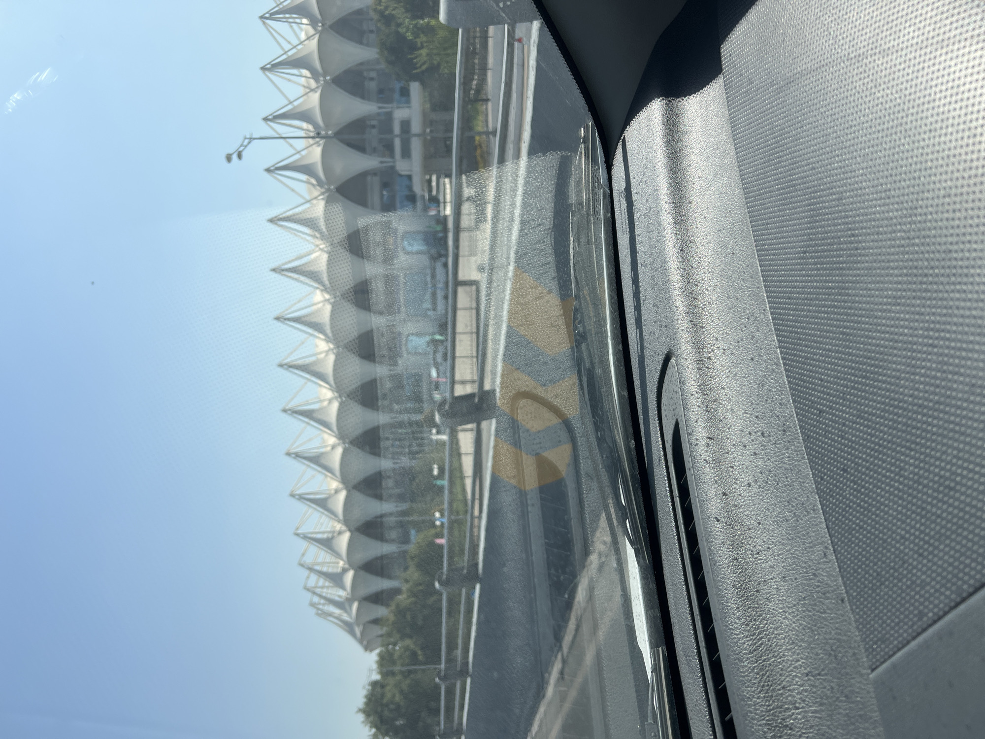 The image shows a view from inside a car looking out through the windshield at a large, modern stadium with a distinctive tent-like roof structure. The stadium is white and appears to be set against a clear blue sky. Trees are visible on either side of the stadium. In the foreground, the car's dashboard is visible, as are the windshield wipers. There are yellow and black striped markings on a barrier in front of the car, indicating a road hazard or lane divider. The overall impression is a sunny day in an urban environment, likely in Tashkent, Uzbekistan, given the context of the request. There are no people clearly visible in the image.