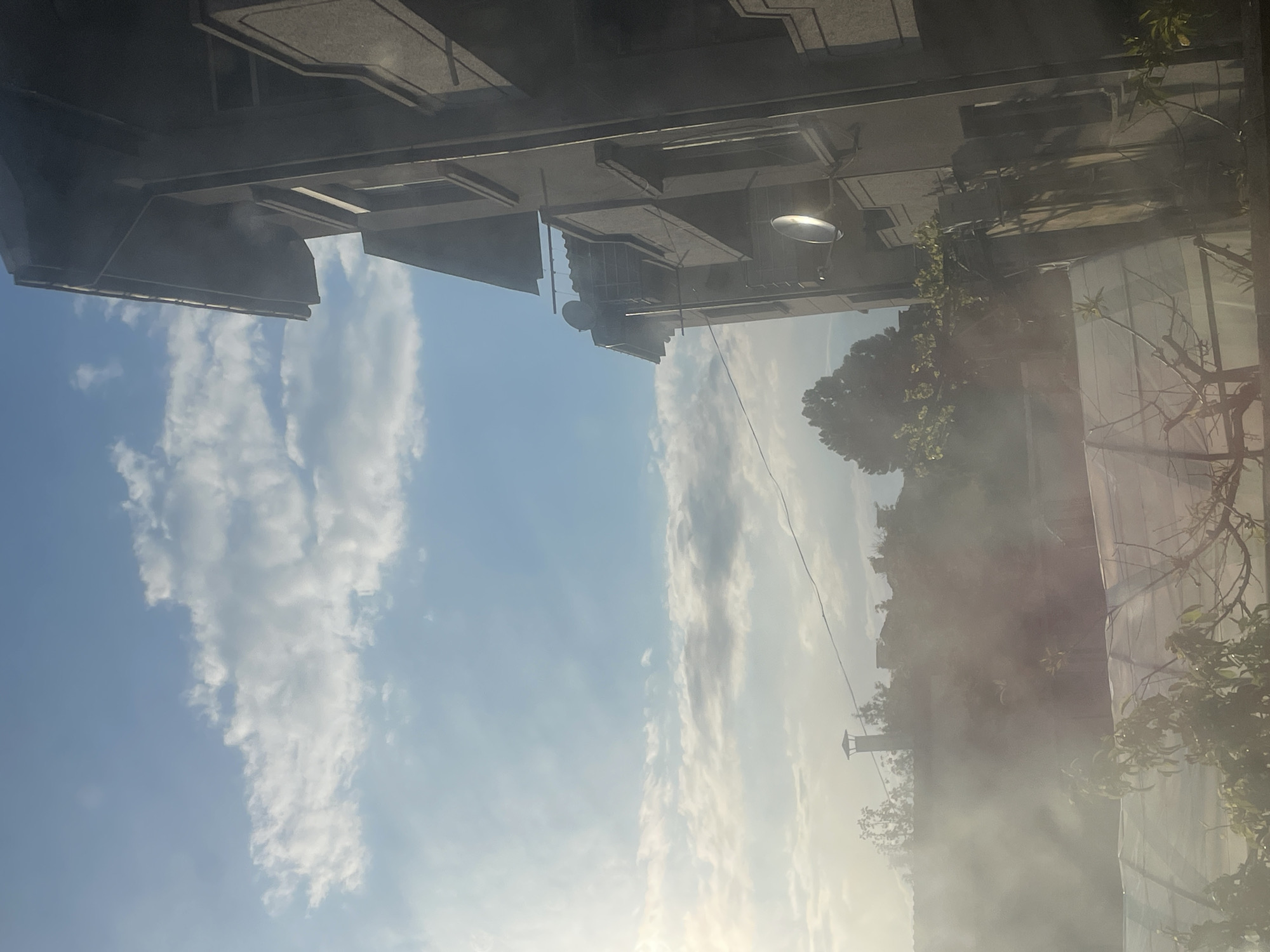 The image is taken looking up at buildings in Harmanli, Bulgaria. The left side shows the corner of a multi-story building with decorative ledges and recessed windows under a bright blue sky with puffy white clouds. In the center is a street lamp attached to another building. On the right side, trees and possibly another building covered in a light haze or smoke are partially visible. A wire stretches from the lamp towards the obscured building. The sun is shining brightly, making the lower portion of the scene appear somewhat washed out.