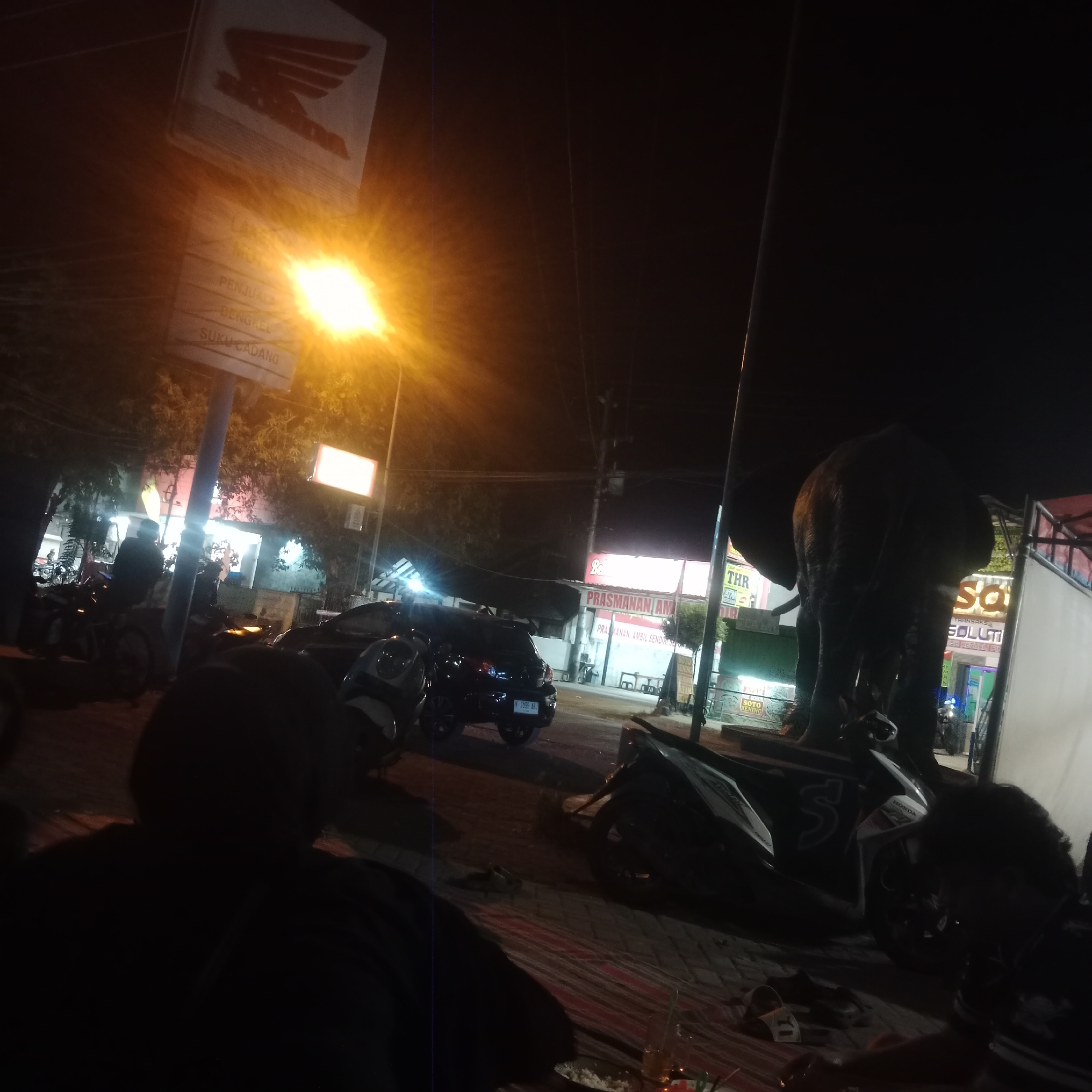 This is a low-light, night-time photograph taken in Pilang Kidul, Indonesia, featuring a street scene with several motorcycles, a car, a large statue of an elephant, and storefronts. In the foreground, there are two individuals, their faces obscured by shadow. One person is seated, facing away from the camera, with a dark hoodie. Another person is also in silhouette, their head and shoulders visible. In the middle ground, a black hatchback car is parked. To the right, a dark, large statue of an elephant is prominently displayed, its back facing the viewer. Behind the elephant, a building with neon signs is visible, one of which reads "PRASMANAN AMBIL SENDIRI." Several motorcycles are parked in front of the establishments. A Honda sign is visible in the upper left portion of the image, above a street sign with Indonesian text, including "PENJUAL," "BENGKEL," and "SUKU CADANG." A bright orange street light illuminates the scene. The ground appears to be paved.