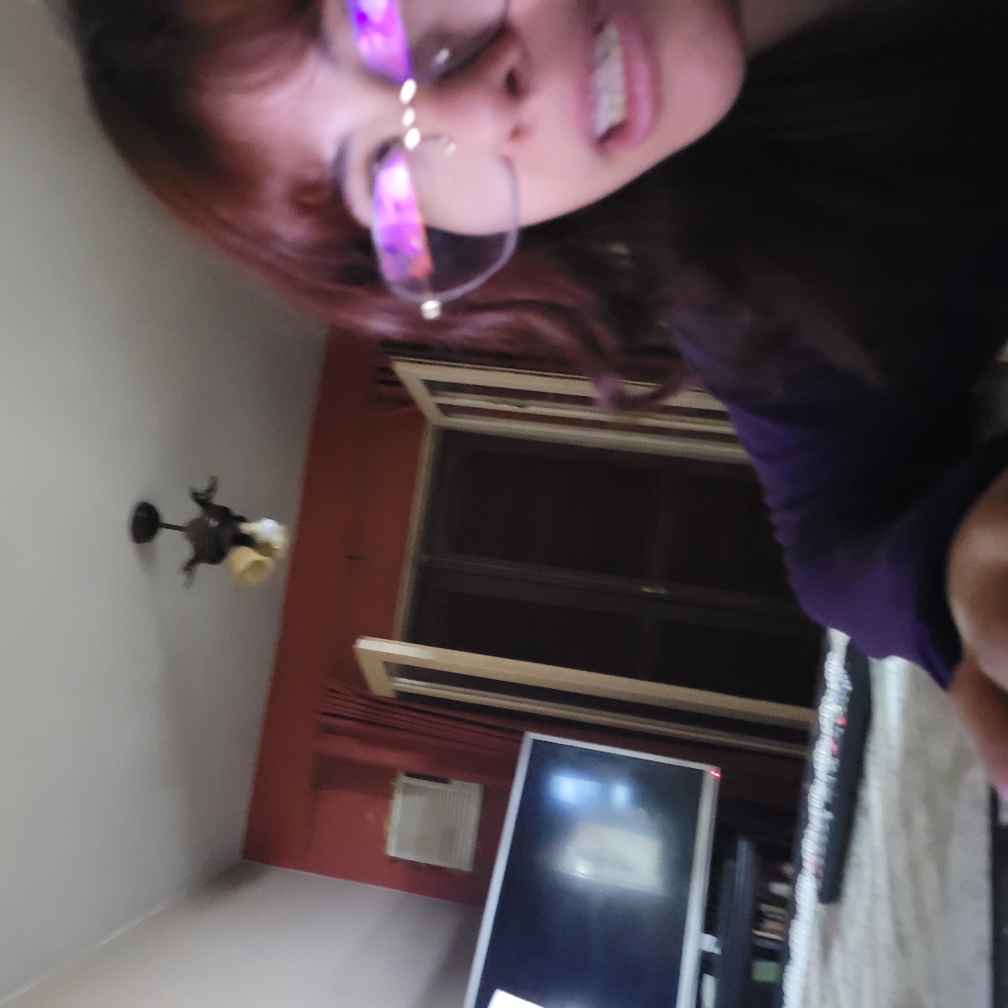 The image presents a tilted view of a room in an unidentified location, likely indoors. A person with auburn-tinted hair, wearing glasses and a purple top, partially occupies the upper portion of the frame. The room features a window with blinds, a television, an air conditioning unit on a red wall, and a ceiling fan. A white, textured surface is visible near the lower right corner, possibly a bed or sofa. The specific city of San Fernando del Valle de Catamarca, Argentina cannot be confirmed from this image.