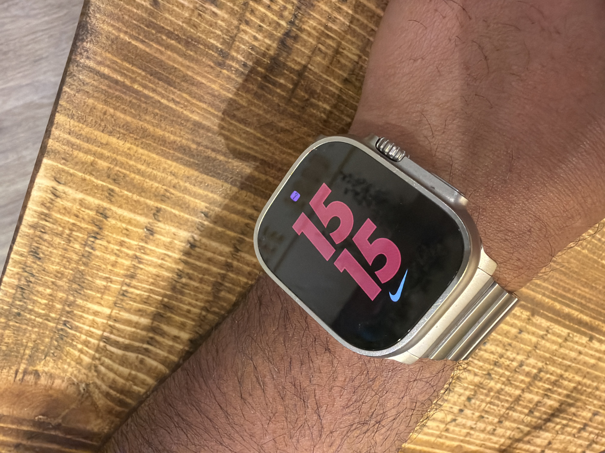 A close-up shot shows a wrist wearing a smartwatch, possibly an Apple Watch Ultra with a silver metal link bracelet. The watch face displays "15 15" in a pinkish-red hue, accompanied by a blue Nike logo. The arm is hairy and appears to be resting on a wooden surface. No specific location in Mole Valley, United Kingdom is discernible from the provided image, as the scene is too close-up and lacks contextual clues.