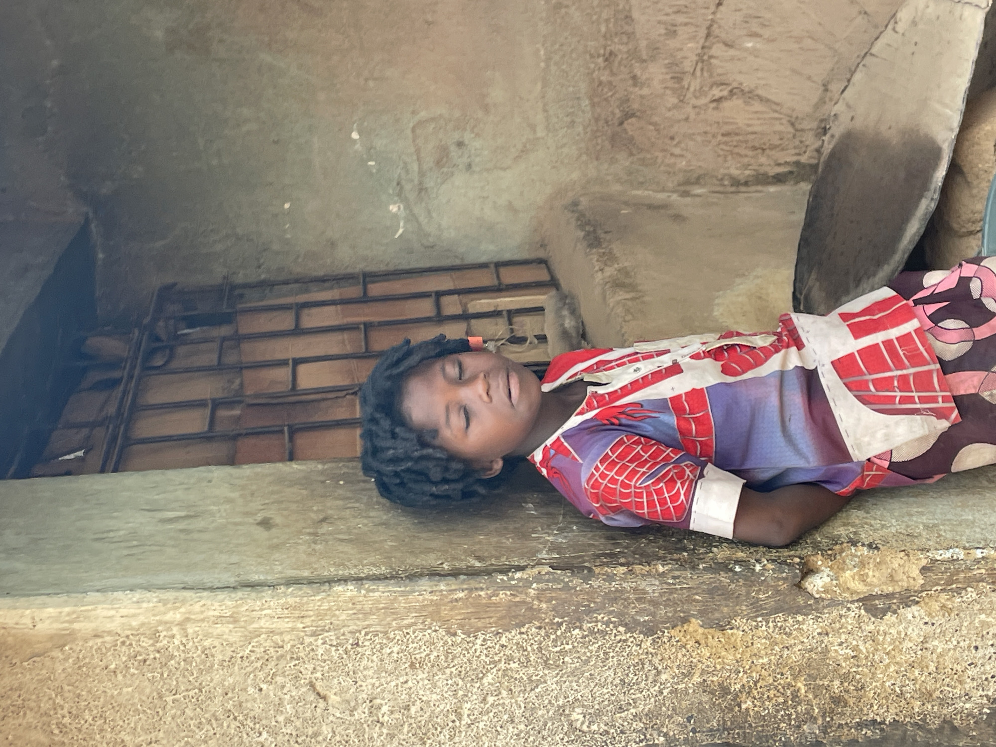 A young Black child with braided hair is sleeping on a ledge. The child is wearing a patterned shirt, with portions showing red spiderweb designs. Behind the child is a barred window. The background environment is rough, aged, and appears to be a building exterior made of textured concrete or plaster. The location is Jos, Nigeria.