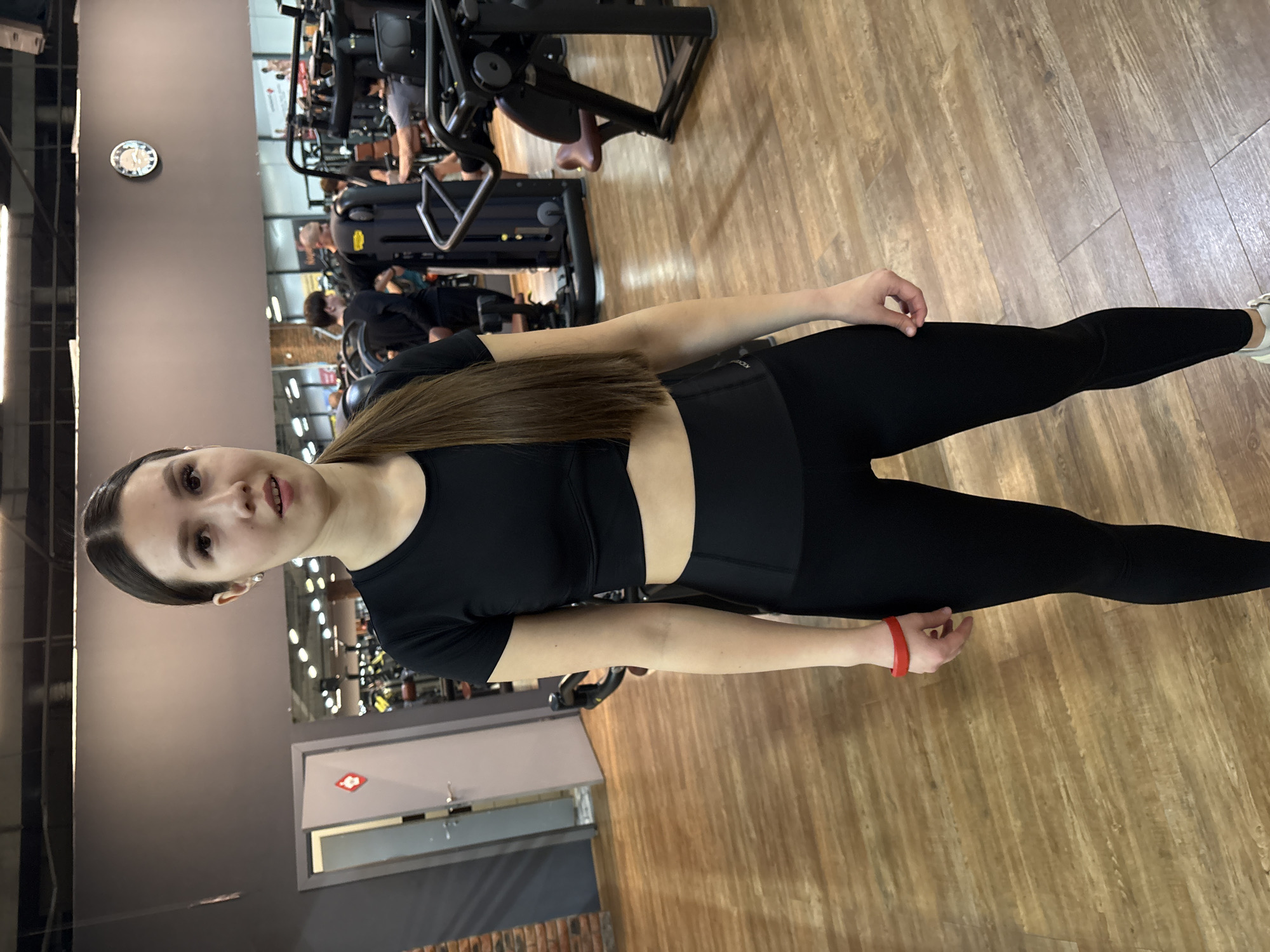 An indoor full-body shot captures a young woman posing in a modern gym in Chaikovsky, Russia. She stands slightly left of center, looking directly at the camera with a soft smile. Her long, dark hair is pulled back, and she wears a black, short-sleeved crop top, black leggings, and white sneakers. A red wristband adorns her right wrist.
The gym features a light wooden laminate floor and dark gray walls. In the background, several other individuals are visible, either exercising on various gym machines or moving around, though their faces are largely obscured or blurred. Gym equipment, including treadmills and weight machines, fills the background. A circular wall clock is mounted on the far wall. To the lower left, a light gray door with a window and a red diamond-shaped sign is visible. The scene suggests an active fitness environment, likely during daytime, with artificial lighting illuminating the space. No readable text is clearly visible other than the clock face.