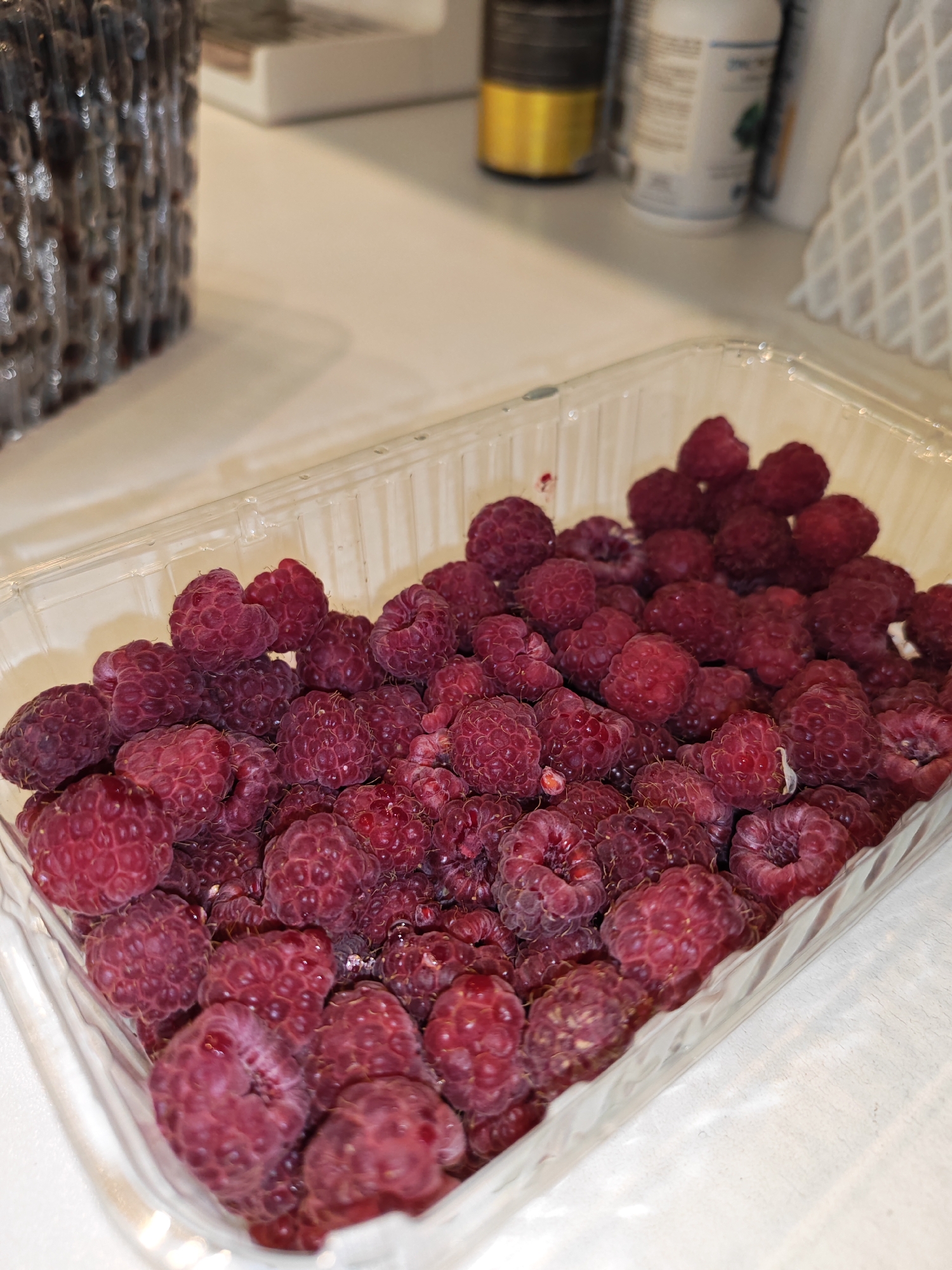 A clear plastic container filled with fresh raspberries is positioned on a white surface. The raspberries are a deep red color and appear plump and ripe. The container is partially filled, with the berries mounded towards the top. The background is blurred, showing what appears to be a kitchen counter with bottles and other containers. There are no people or animals visible in the image. The lighting is soft and natural, suggesting an indoor setting. The image does not provide any information about its location in Bataysk, Russia.