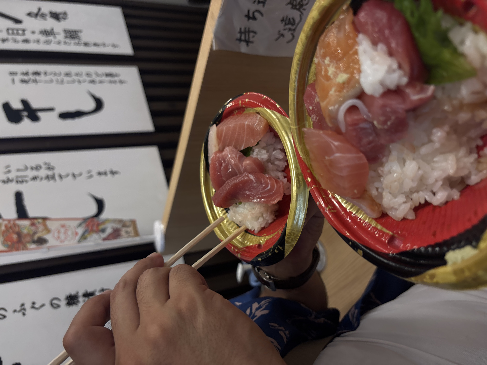 The image captures a person's hands holding chopsticks, engaged in eating a meal in Kanazawa, Japan. Two small, decorative bowls filled with kaisendon or chirashi sushi are prominent, featuring sushi rice topped with various slices of raw fish, including vibrant red tuna, pink salmon, and white fish. A touch of green garnish is visible in the bowl further from the camera. The bowls themselves are ornate, displaying traditional red, black, and gold designs.
The person is dressed in a dark blue garment with a subtle pattern, and a watch is visible on their left wrist as they skillfully maneuver the wooden chopsticks. The activity is the simple act of enjoying a fresh seafood dish, likely at a restaurant, food stall, or market.
In the background, several signs with handwritten Japanese text are visible, though partially out of focus. These signs offer notable location cues. Deciphered text includes references to "いしる" (ishiru), a regional fish sauce originating from the Noto Peninsula, and "ふぐの糠漬け" (fugu no nukazuke), fermented pufferfish in rice bran, both well-known local specialties of Ishikawa Prefecture, where Kanazawa is situated. Other fragments include "持ち主" (owner) and "ご遠慮" (please refrain), possibly part of a polite request or instruction. The indoor setting, with wooden panels visible, and the specific regional food items mentioned on the signs, strongly confirm the location to be Kanazawa, Japan, highlighting an authentic culinary experience.