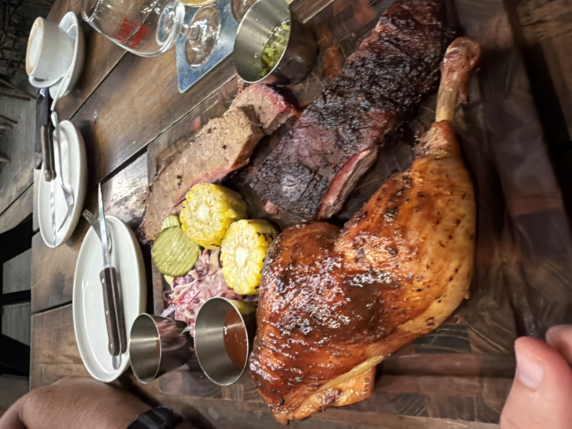 The image depicts a wooden serving platter laden with food, likely a meat platter, and dining accoutrements. The platter includes a large turkey leg with a shiny, glazed surface, a section of beef ribs with a darkened, slightly charred exterior, sliced brisket, corn on the cob, pickles, and coleslaw. There are also several small metal ramekins, one containing a green sauce and another a red sauce. A stack of plates with silverware is visible to the left, along with a coffee cup and what appears to be a beer sampling tray with multiple glasses. The lighting suggests an indoor setting, possibly a restaurant. The location is identified as Nha Trang Ward, Vietnam. A person's hand and arm can be seen in the corner of the image, suggesting a person is seated at the table.