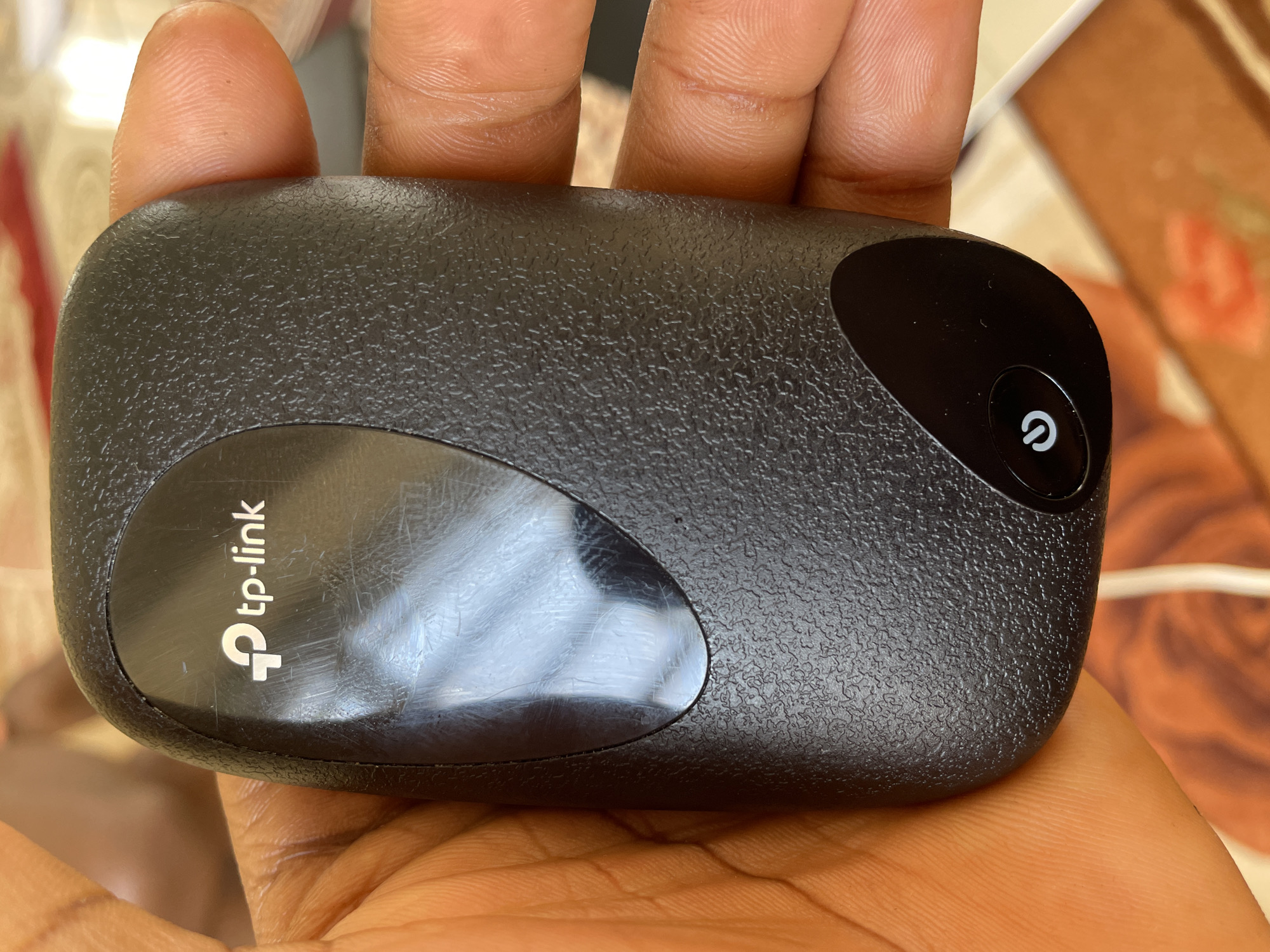 A hand holds a black TP-Link portable Wi-Fi device. The device has a textured surface and a glossy black oval on the left side with the TP-Link logo in silver. Another glossy black oval on the right side contains a white power button icon. The background is out of focus and appears to be an indoor setting with warm-toned furniture or decorations. The lighting suggests it is daytime. The image is a close-up shot of the device, with fingers holding it from underneath. The context indicates the location is Wai A, Nigeria.