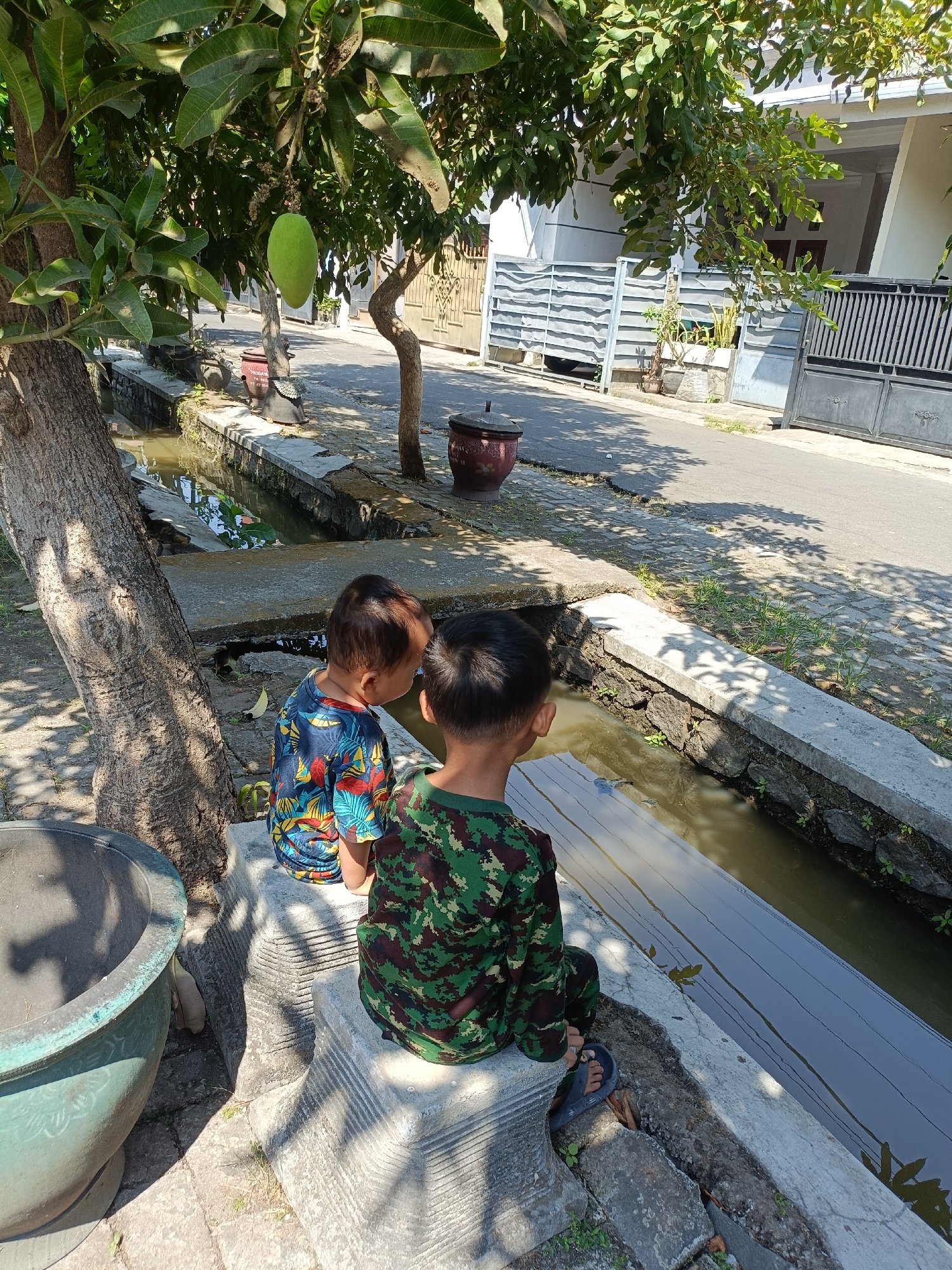 Two young boys are sitting on a stone bench beside a narrow, shallow canal filled with murky water. The boy on the left wears a colorful short-sleeved shirt with a floral pattern. The boy on the right wears a camouflage-patterned shirt and pants. They are both facing the canal. In the background, there is a street with a house and a metal fence. A mango tree with a single green mango hanging from a branch is visible to the left of the frame. The scene appears to be an outdoor residential area, likely in a tropical or subtropical climate given the foliage and the presence of a mango tree. The overall lighting suggests it is daytime with bright sunlight casting shadows. The location is identified as Kediri City, Indonesia.