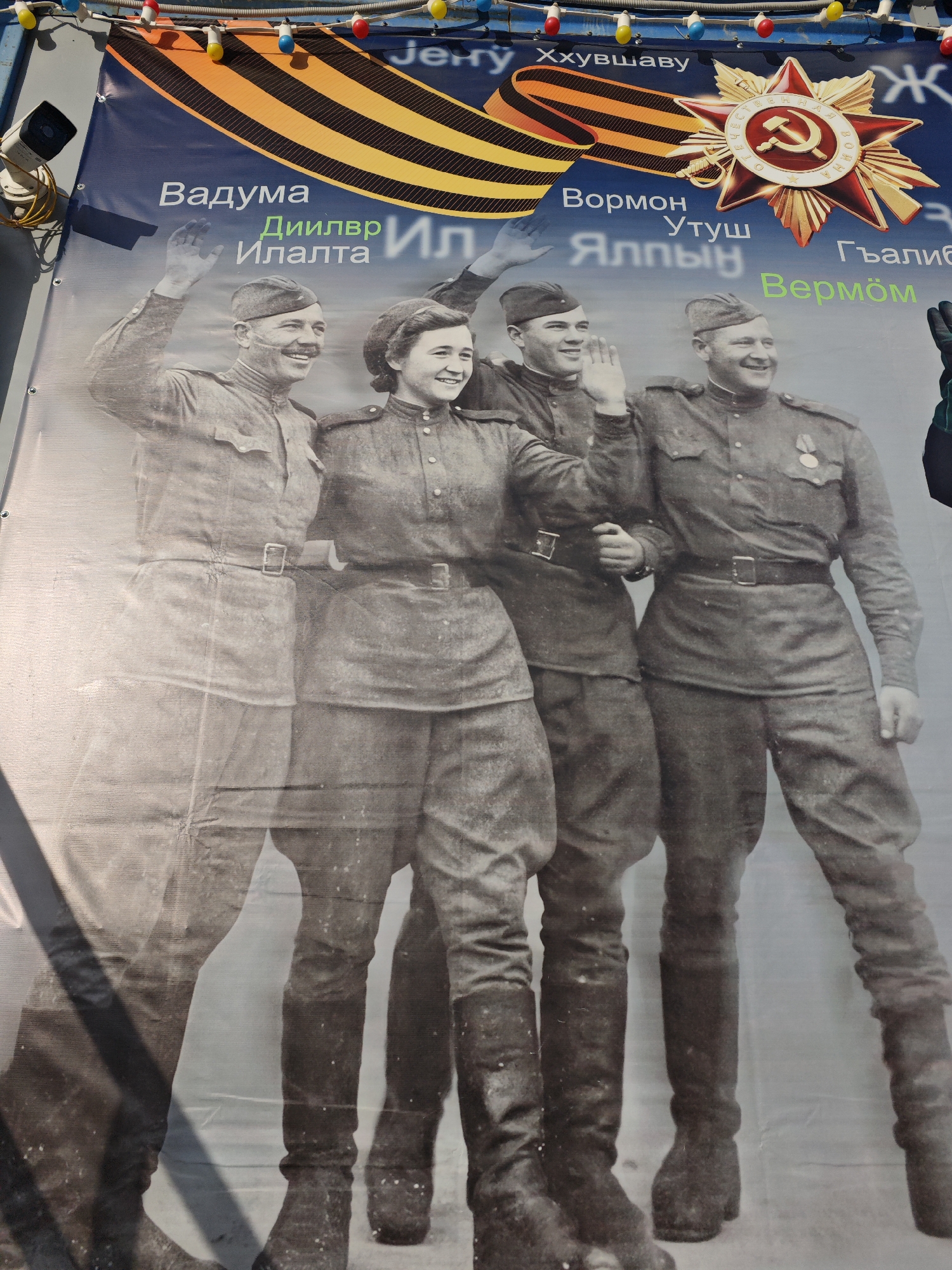 A banner in an unknown city in Russia features a black-and-white photograph of four smiling soldiers in uniform, waving. Above the photo is a ribbon with black and orange stripes and a star emblem with a hammer and sickle. Text is written in a foreign language above the image and next to the soldiers' heads. A string of decorative lights is visible at the top of the banner.