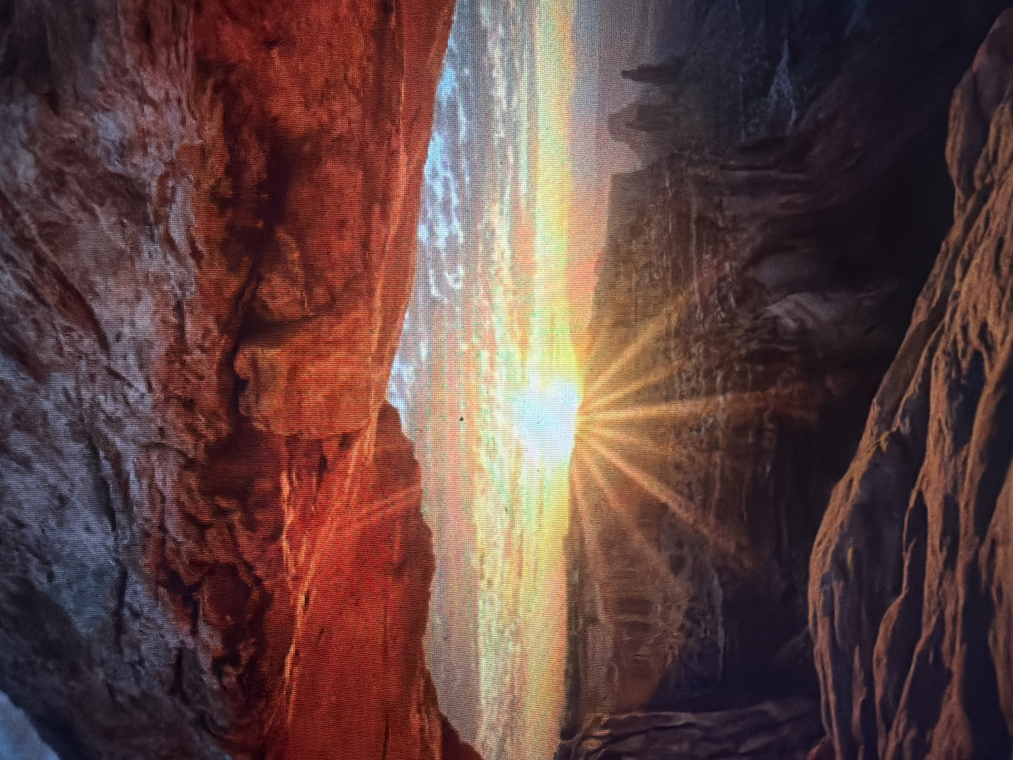 The image depicts a scenic view through a natural rock arch. The arch, predominantly red and textured, frames a vibrant sunset over a canyon landscape. The sun, a bright orb with radiating beams, is positioned near the horizon, casting a warm glow across the sky and illuminating the distant rock formations and the canyon's depths. The sky is filled with a mixture of colorful clouds, ranging from orange and yellow near the sun to cooler blues and grays further away. The foreground shows rugged, rocky terrain, possibly part of the arch itself or the surrounding landscape. The overall scene is one of natural beauty and dramatic lighting, characteristic of a sunset in a desert or canyon environment. The image does not contain any elements that suggest it is located in Baan Mae Nam, Thailand.