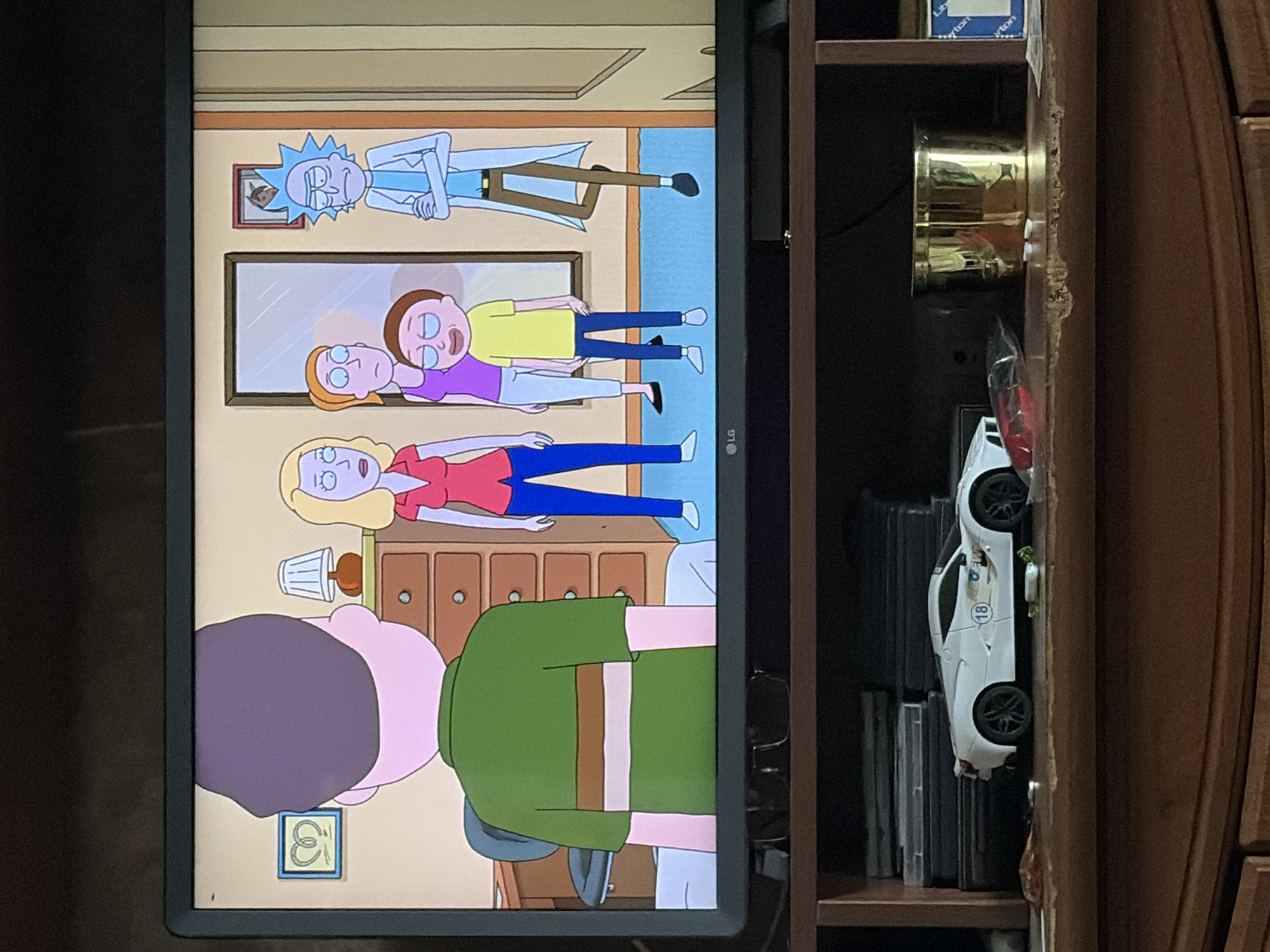 The image shows a television screen displaying an animated scene from the show Rick and Morty. In the foreground, a person wearing a green shirt with brown and white stripes is facing away from the viewer. To the right of this person, on the TV screen, are three other animated characters: a woman with blonde hair wearing a red shirt and blue pants, a woman with orange hair wearing a purple top and white pants, and a young boy with brown hair wearing a yellow shirt and blue pants. To the far right is an animated character resembling Rick Sanchez in his lab coat. The scene appears to be set indoors, with a dresser and a doorway visible in the background. Below the television, on a wooden shelf, is a white toy sports car with the number "18" on its side, and a gold cylindrical object. The shelf appears cluttered with other items, including what looks like a stack of DVDs or game cases. The setting is a domestic interior, likely a living room or entertainment area.