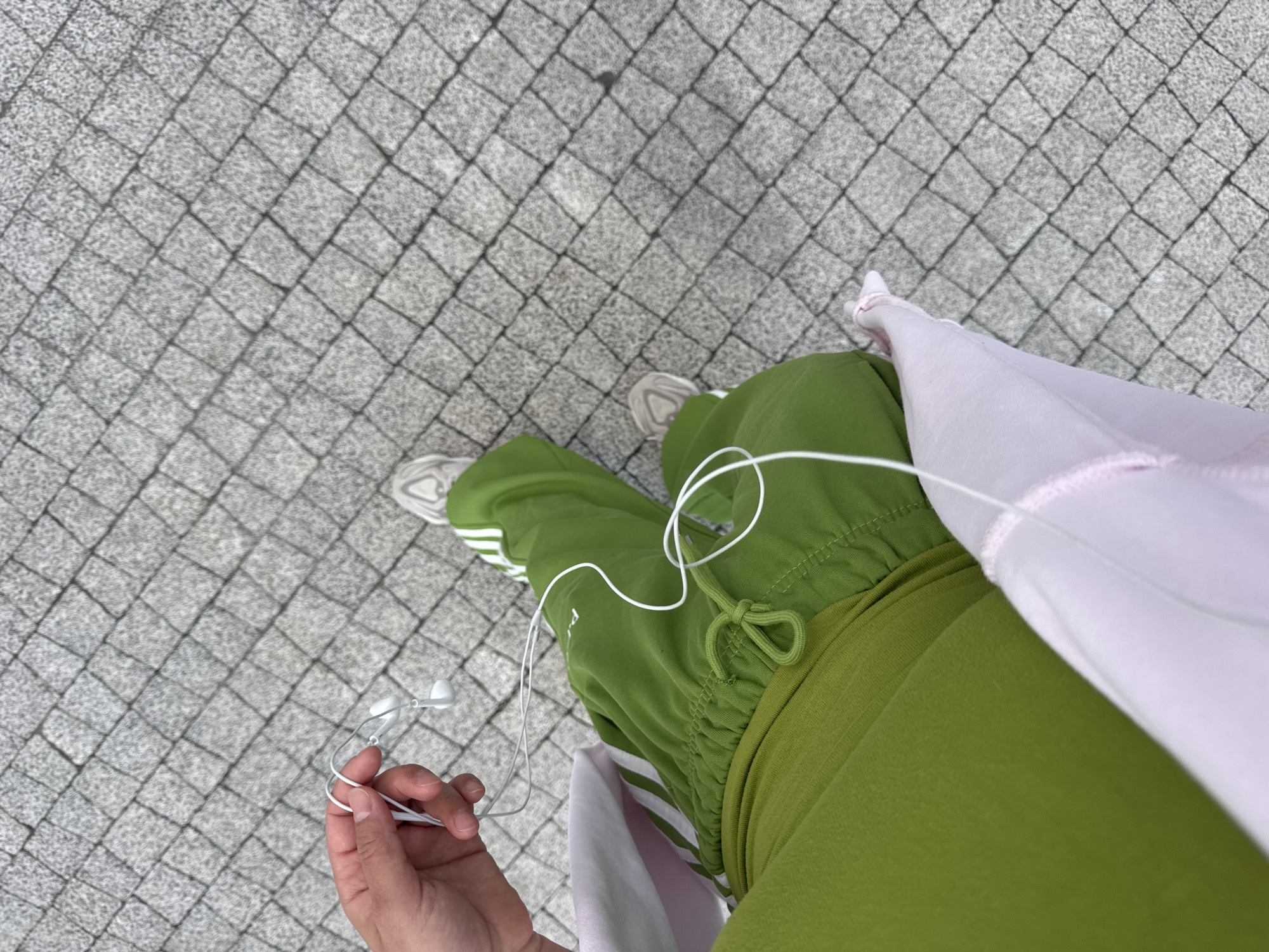 A first-person, overhead view depicts a person standing on a gray cobblestone pavement. The person is wearing green Adidas track pants with white stripes and white sneakers. They are holding white wired earphones in their left hand. A pink and white garment is partially visible on their right side. The location is inferred to be Kazan, Russia.
