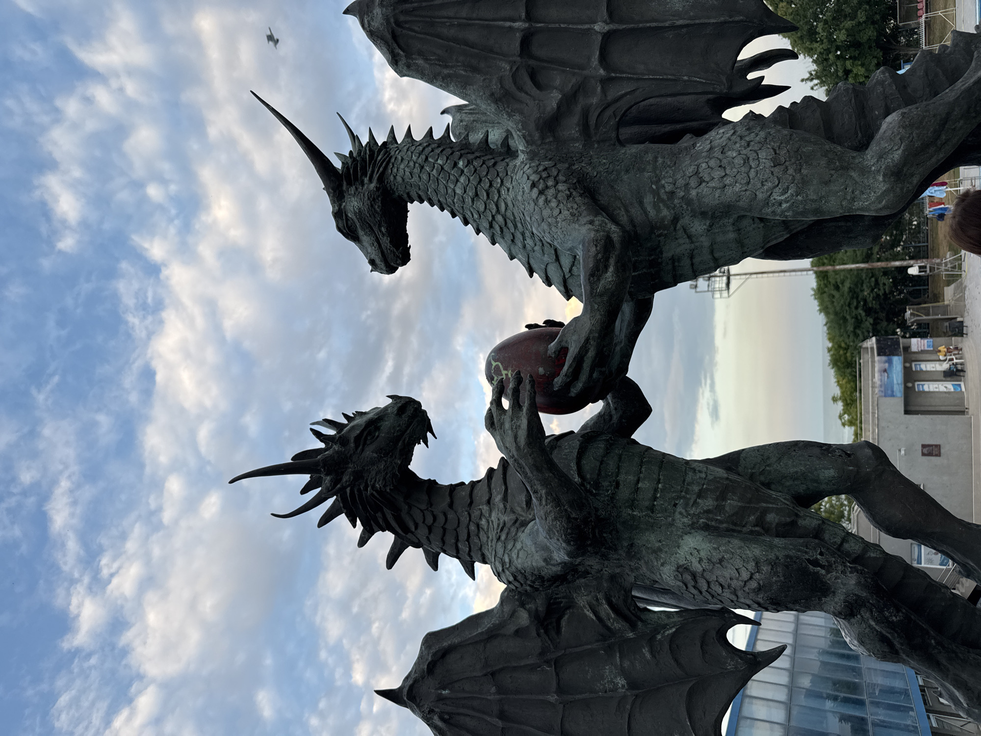 The image shows a sculpture of two dragons against a cloudy sky. The dragons are made of dark metal, likely bronze, and are depicted in a dynamic pose, with one dragon holding a red, spherical object. The sculpture appears to be outdoors, with trees and buildings visible in the background, suggesting a park or public space. The sky is partially overcast with scattered clouds, indicating it could be daytime, possibly late afternoon or early evening given the soft lighting. The overall mood of the image is dramatic and mythical, due to the subject matter of dragons. There is no visible text in the image. The location context provided is Varna, Bulgaria.