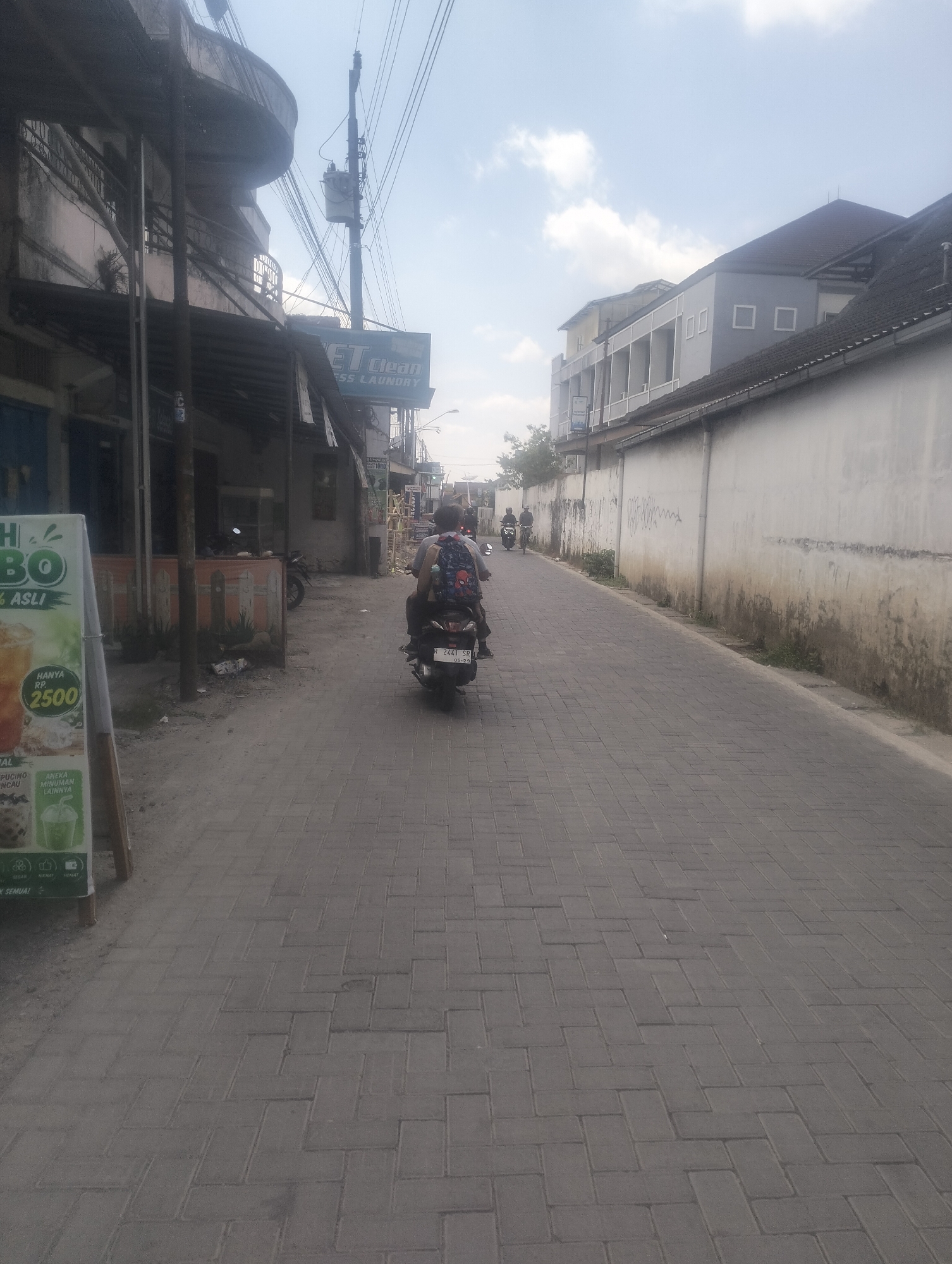 The image shows a street scene in Karangsoka, Indonesia. A paved road with brick pavers runs through the middle of the frame, lined with buildings on either side. On the left, a building with balconies and awnings appears to be a commercial establishment, possibly a shop or restaurant, with a sign that reads "HANYA RP 2500" and mentions drinks. Further down the street on the left, a utility pole with numerous wires is visible. On the right side of the street, a long, white wall with some discoloration and graffiti stretches into the distance. Behind the wall, modern buildings with multiple stories and gray facades are visible.
In the foreground, a person is riding a motorcycle with a passenger on the back, both wearing backpacks. The motorcycle has a license plate that reads "R 2441 SR." In the distance, other motorcycles can be seen on the road, suggesting moderate traffic. The sky is cloudy, and the overall lighting suggests it is daytime. The street appears to be a commercial or residential area within the city.