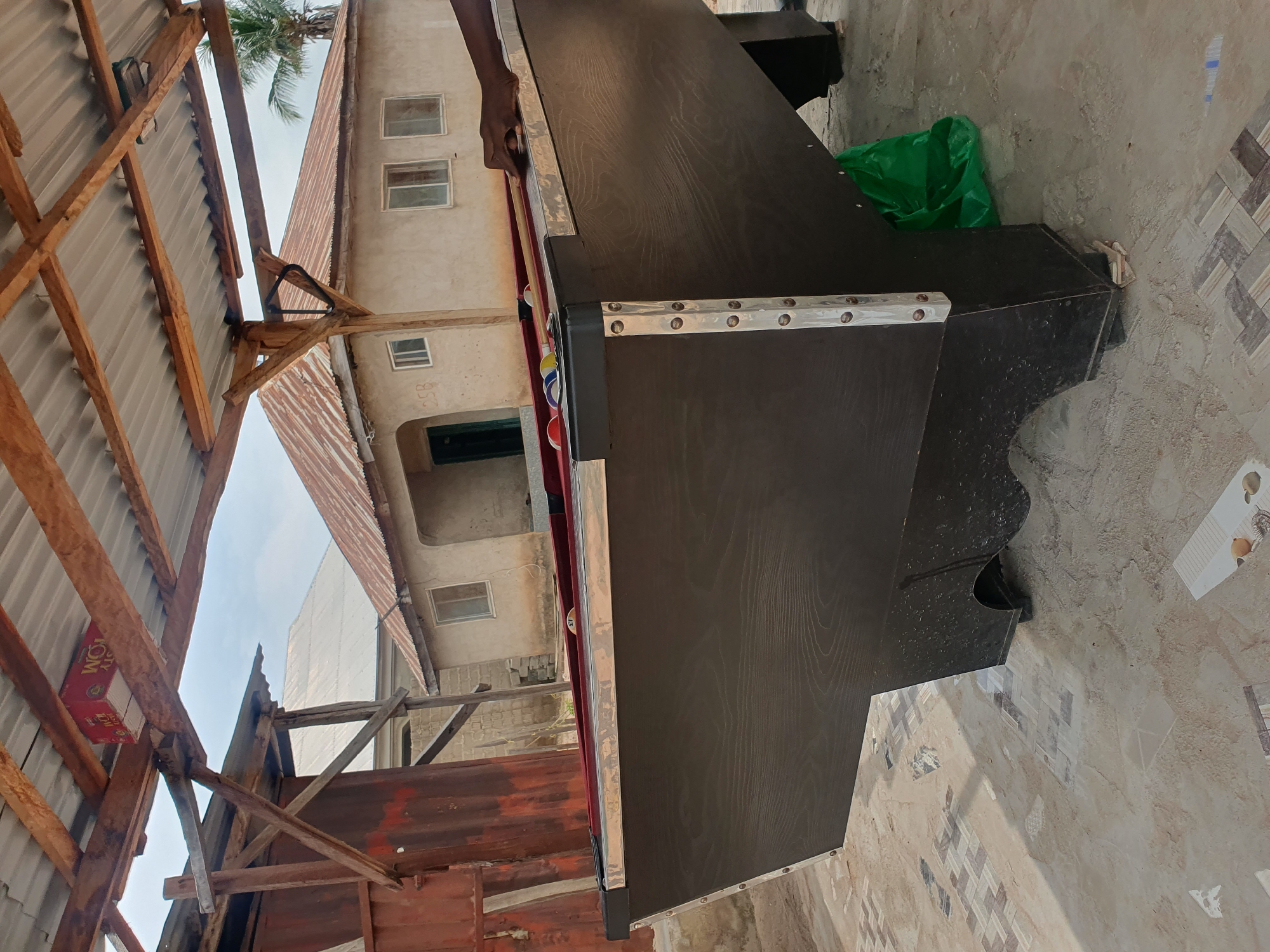 In Lokoja, Nigeria, an outdoor scene depicts a billiard table under a makeshift corrugated metal roof supported by wooden beams. The table is dark wood with a red felt surface, and billiard balls are visible on it. A person's arm and hand are shown reaching out towards the table, holding a cue stick. In the background, a light-colored building with windows and a corrugated metal roof stands under a cloudy sky, with a palm tree visible to the right. The ground is paved with irregular tiles and scattered debris. A green plastic bag is placed under the billiard table.