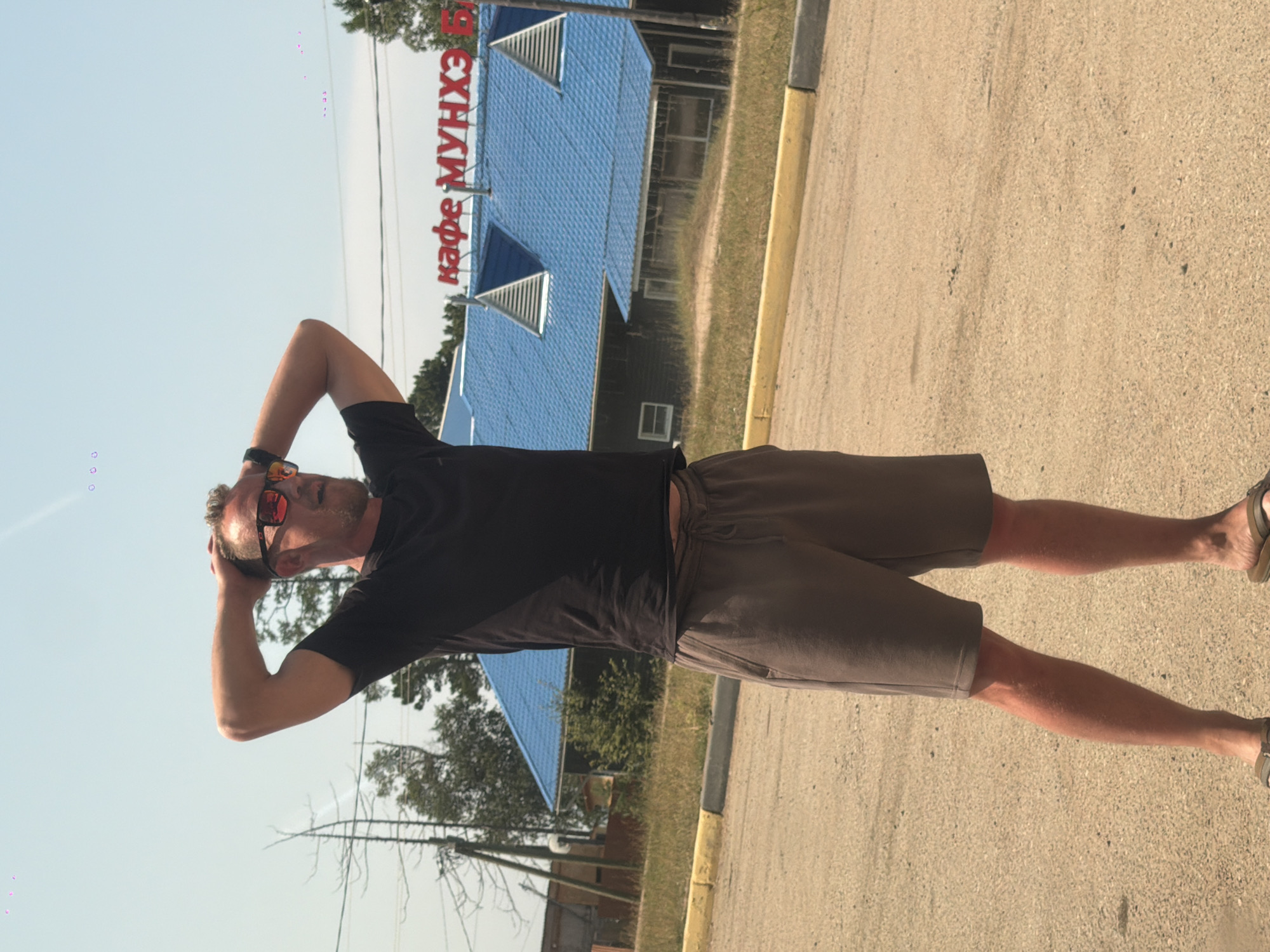 A Caucasian man with blond hair and a beard stands in a dirt lot, his hands behind his head. He is wearing a black t-shirt, brown shorts, and sandals, and sunglasses. Behind him, a building with a blue roof has red lettering that reads "кафе МУНХЭ Б" which translates to "Cafe Munhэ B". The building appears to be a cafe or restaurant. The scene is outdoors on a sunny day with a clear blue sky. The location is in Turka, Russia.