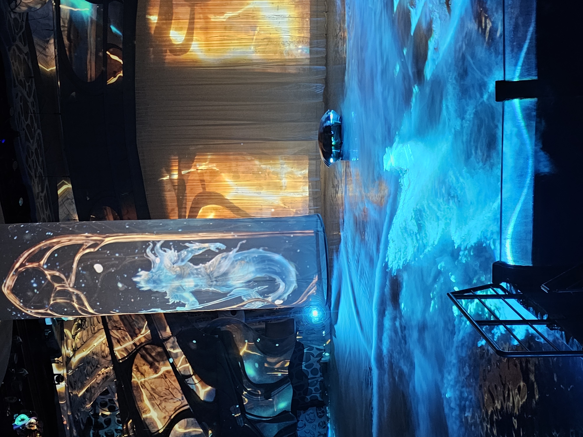 The image captures an immersive art installation, likely in a darkened, enclosed space. In the foreground, a horizontal display case features a stylized image of a light blue, mystical figure within a decorative, cage-like structure. This is reflected on the surrounding walls.
To the right of the case, a vertical projection displays an abstract, flowing pattern dominated by vibrant blue and white hues, resembling a waterfall or a dynamic water effect.
Behind the display case and to the left, the architecture suggests a gothic or ornate design, with dark structures and glimpses of what might be more projections or light displays in shades of orange and brown.
Overall, the scene conveys a sense of an artistic and technologically advanced exhibition. The location cannot be definitively identified as Moscow, Russia from the image.