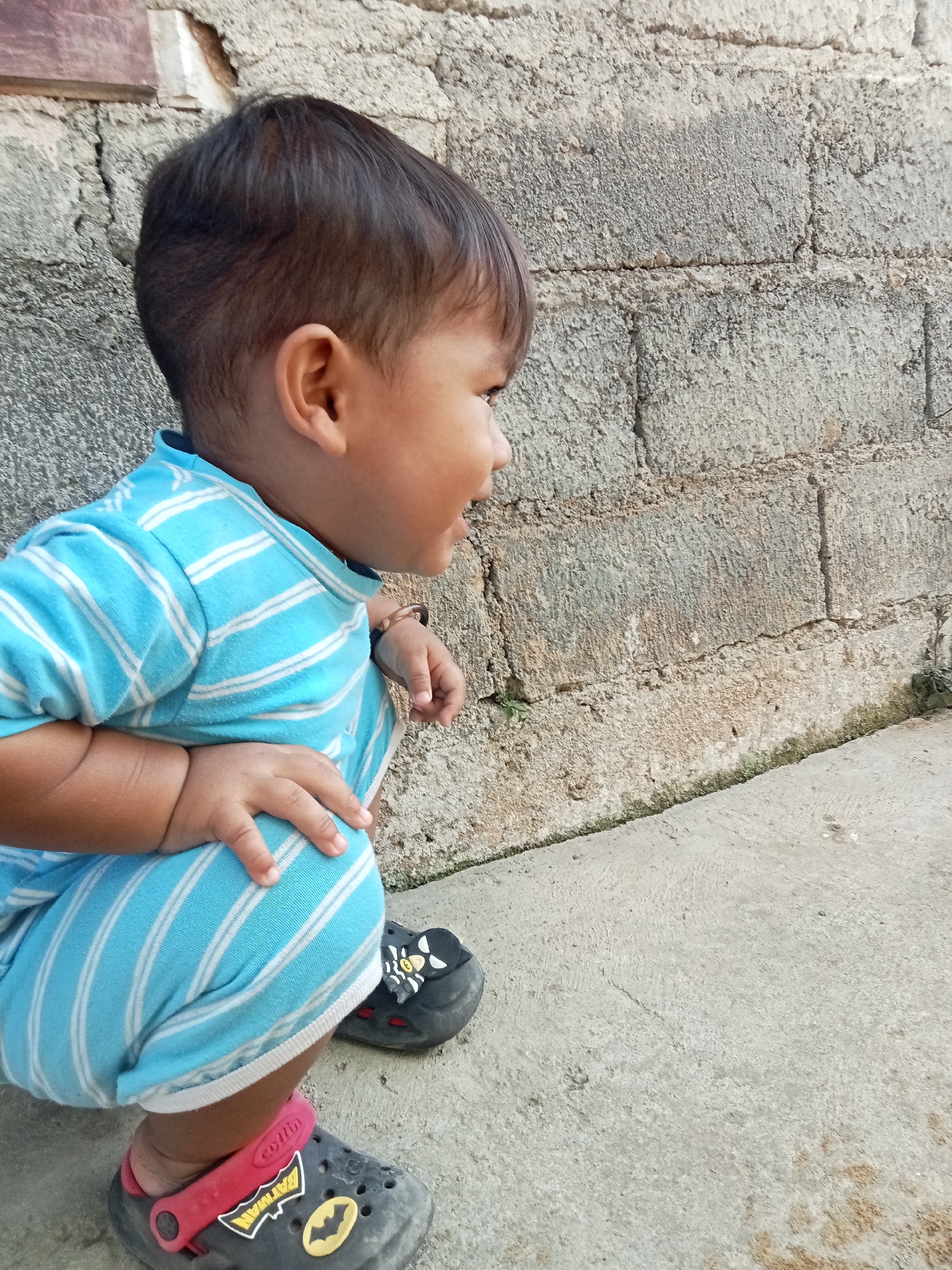 A young child with dark hair and light skin kneels on a concrete surface. The child wears a blue and white striped shirt and pants. The child's feet are clad in dark gray clogs adorned with Batman logos. Behind the child is a rough, textured gray brick wall. The child is looking to the right with an open mouth. The environment appears to be outdoors, possibly in the city of Pancurwening, Indonesia, given the weathered brick wall and concrete ground, common features in residential areas of the region.