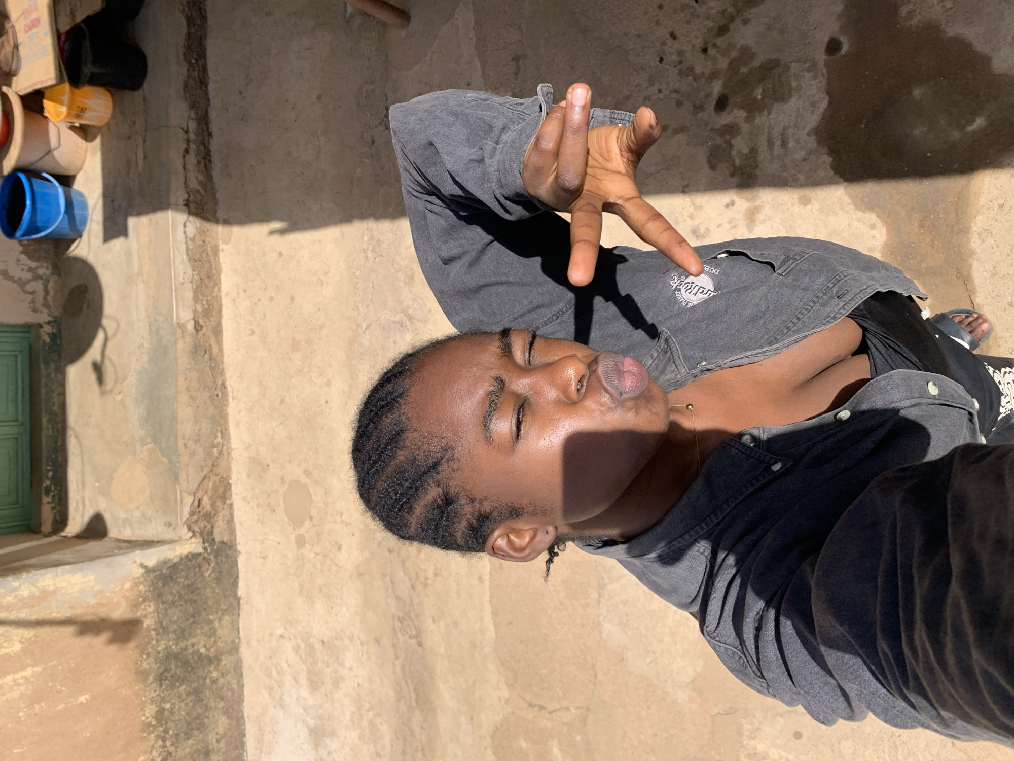 The image depicts a person posing outdoors on what appears to be a paved surface. The individual has dark skin and braided hair and is wearing a gray denim jacket over a black top. They are making a hand gesture, possibly a peace sign or a similar symbol, with one hand raised toward the camera. The person is looking into the sunlight with their eyes closed.
In the background, there are several buckets, containers, and a possible cardboard box piled against a wall. A green door is also visible. The setting appears to be an urban environment. Based on the prompt's location hint, this location is in Rafin Par, Nigeria. The person appears to be standing on a paved area, potentially a courtyard or walkway. Shadows are present, indicating strong sunlight.