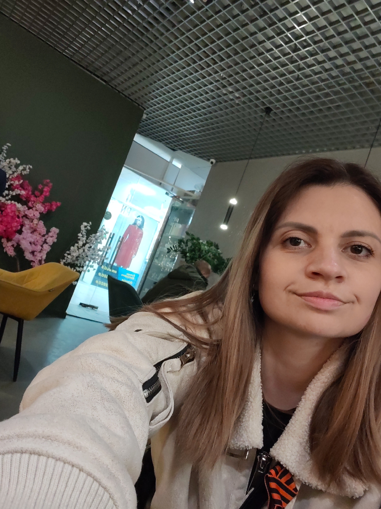 A woman poses for a selfie in what appears to be a public space with a modern interior. The woman has light brown hair and is wearing a white jacket with a fuzzy collar. Behind her, there's a glimpse of a retail storefront with a mannequin displaying a red garment. A yellow chair and pink flowers decorate the space. The ceiling is a grid-like structure, suggesting a commercial or public building. There is no direct information about the location being in Izhevsk, Russia.