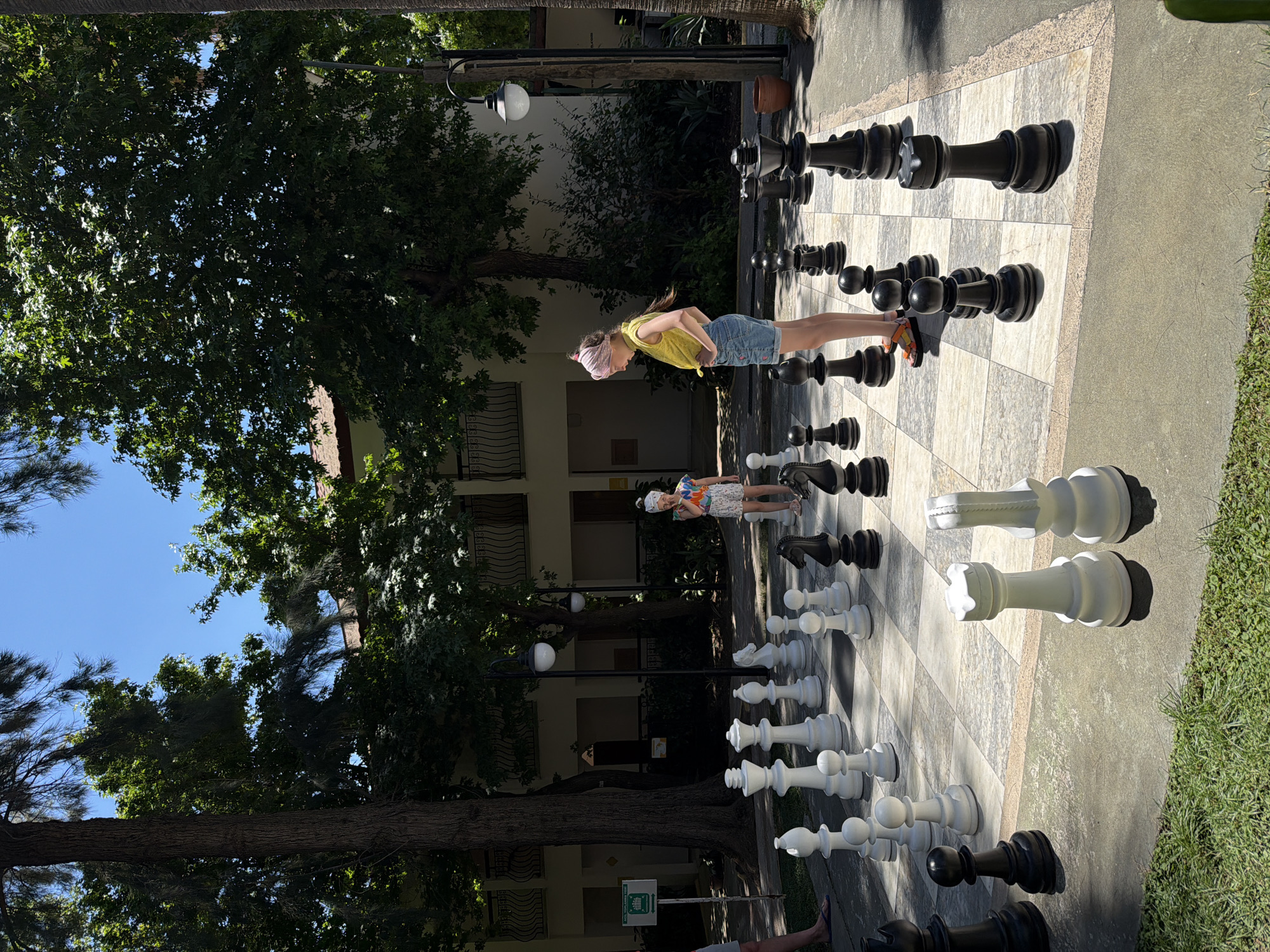 A sideways-oriented outdoor shot features a giant chessboard with chess pieces, where two girls are standing on the board. The girl nearest the camera is wearing a yellow shirt and denim shorts with a pink headband. The other girl, further away, is wearing a light-colored dress with a white hat. The chessboard and pieces are surrounded by trees, and a building is visible in the background. The location is Fethiye, Turkey.