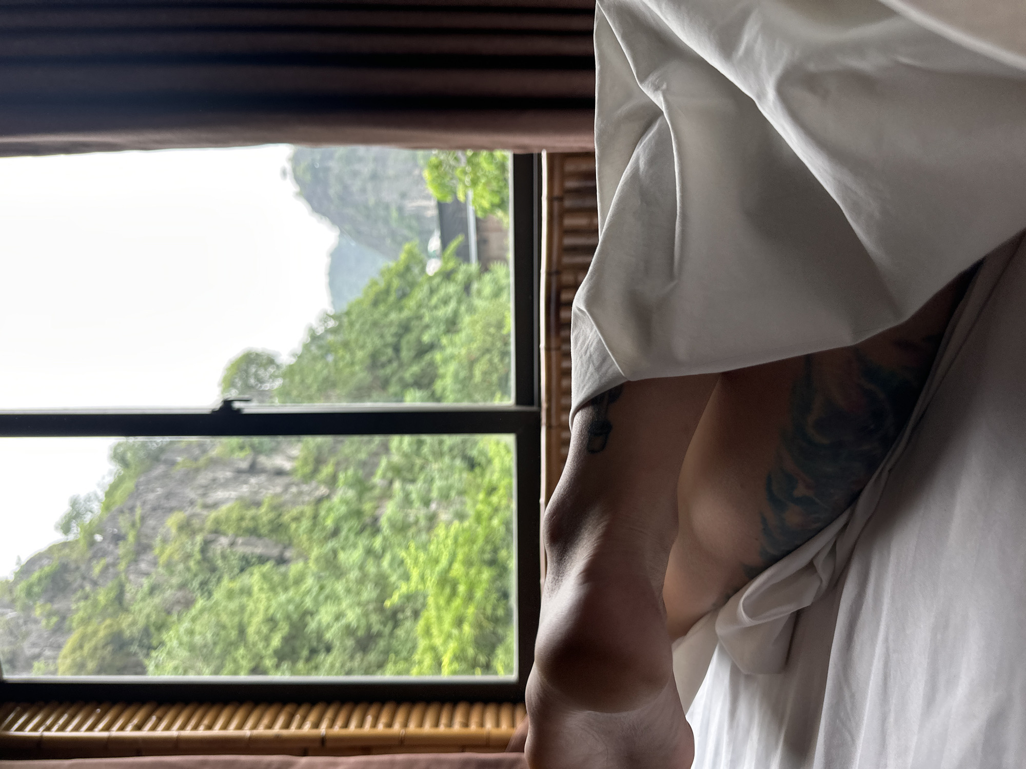 A person lies in a bed in a room with a large window overlooking a lush green mountainous landscape. The room has wooden blinds on the left and a dark brown curtain on the right. The bed is covered with white sheets, and a portion of the person's legs, partially covered by a white duvet, is visible. The person has tattoos on their legs. The view outside the window shows a dense forest covering steep, rocky cliffs, with some buildings visible in the distance. The sky is overcast. The scene appears to be in Nam Hoa Lư Ward, Ninh Bình Province, Vietnam, known for its karst topography.
