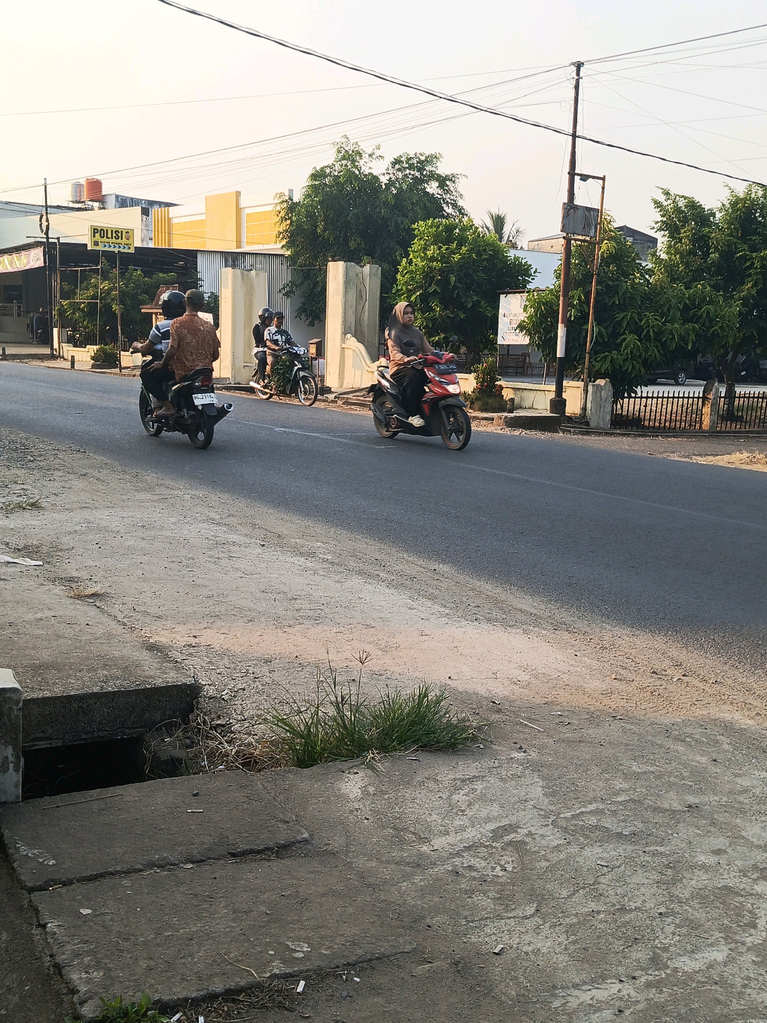 In the city of Talang Ubi, Indonesia, a road scene unfolds under a clear sky. A police station with a yellow and white facade and a prominent "POLISI" sign is visible on the left. Several individuals are operating motorcycles on the paved road. Two people are riding on a dark motorcycle, with the rear passenger wearing a patterned orange shirt. Another motorcycle is carrying a rider and a passenger, also wearing helmets. Further down the road, a person wearing a hijab is riding a red motorcycle. The environment includes trees lining the street, a metal fence, and utility poles with overhead wires. The foreground shows a concrete drain cover and surrounding rough ground with scattered debris. The time of day appears to be late afternoon, as indicated by the warm lighting and long shadows.