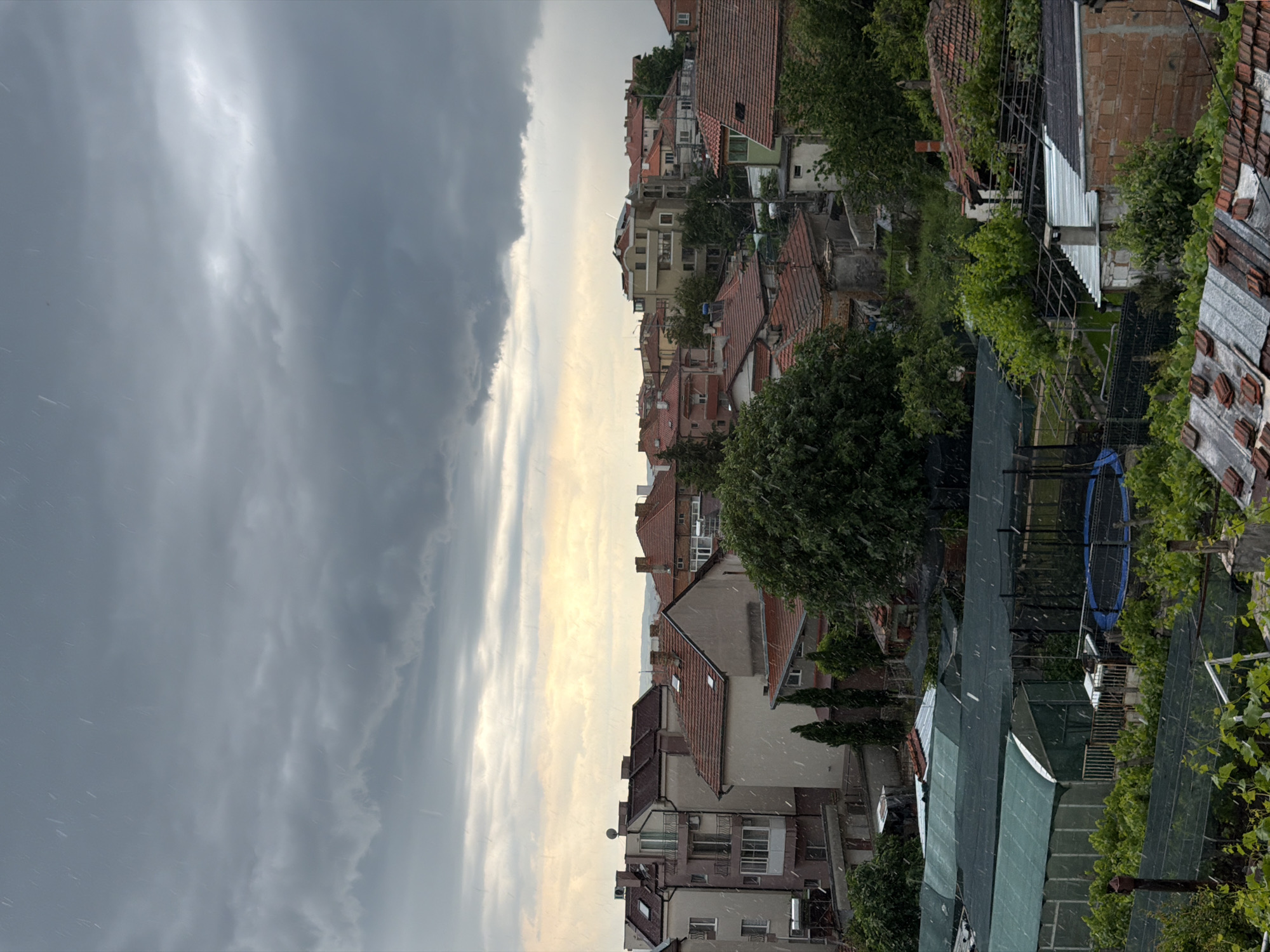 The image shows a view overlooking rooftops in what appears to be Burgas, Bulgaria, based on the architectural style. A storm is in progress, as evidenced by the heavy, grey clouds dominating the sky, and the visible streaks of rain or hail falling. The foreground features residential rooftops, some with traditional red tile roofs, interspersed with green vegetation, including trees and vines. A blue trampoline is visible. The buildings in the distance vary in height and style, typical of a city with diverse architectural periods. The weather seems harsh, suggesting a sudden downpour affecting the urban landscape.
