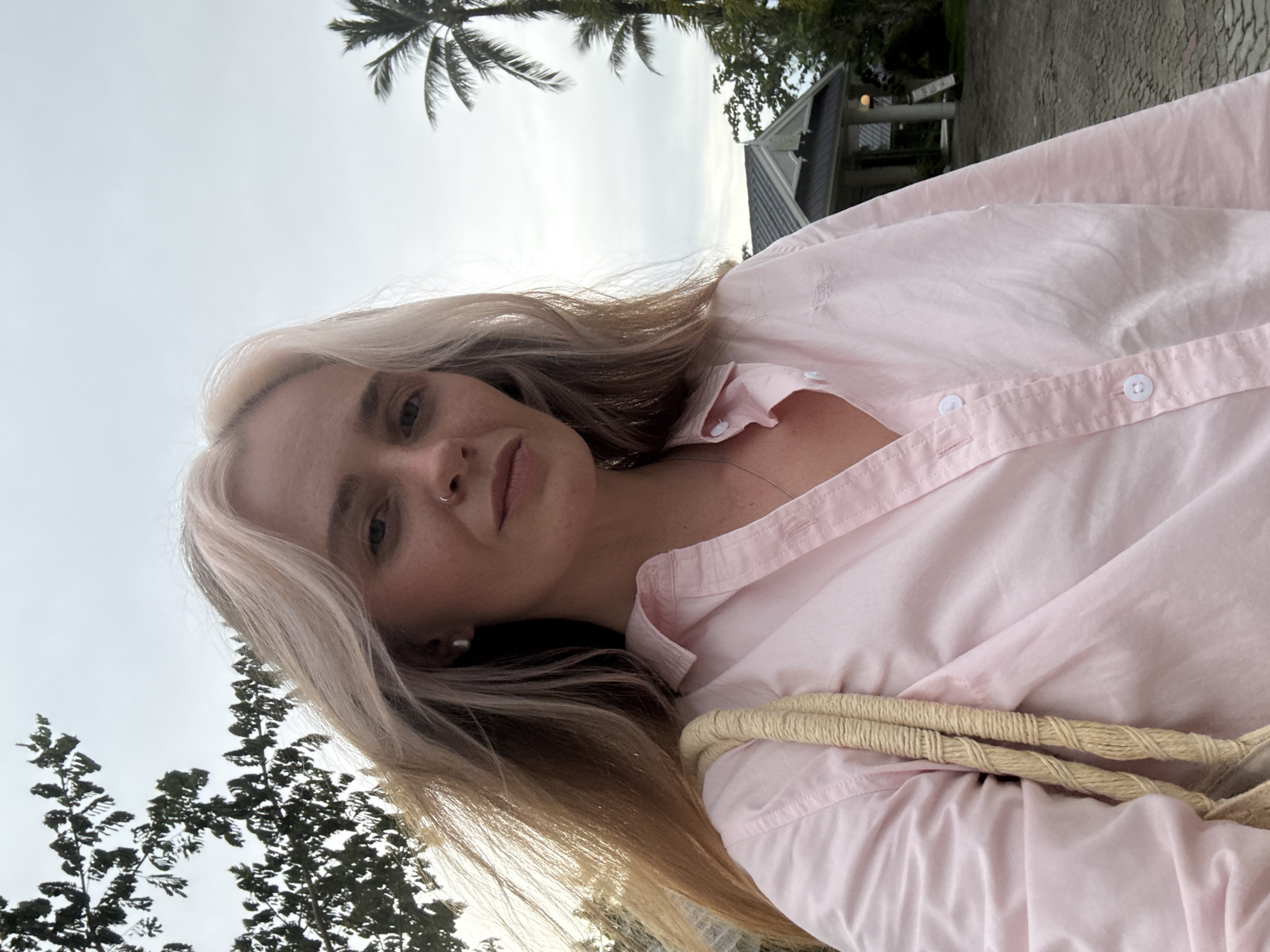 A fair-skinned woman with blonde hair, partially obscured by wind, is pictured from the chest up. She is wearing a pink collared shirt and a tan rope bag over her left shoulder. A nose ring is visible. In the background, a building with a dark roof and a light-colored facade is visible, along with palm trees and foliage. The scene appears to be outdoors, possibly in a tropical setting like Choeng Thale, Thailand, given the presence of palm trees. The lighting suggests it is either early morning or late afternoon.