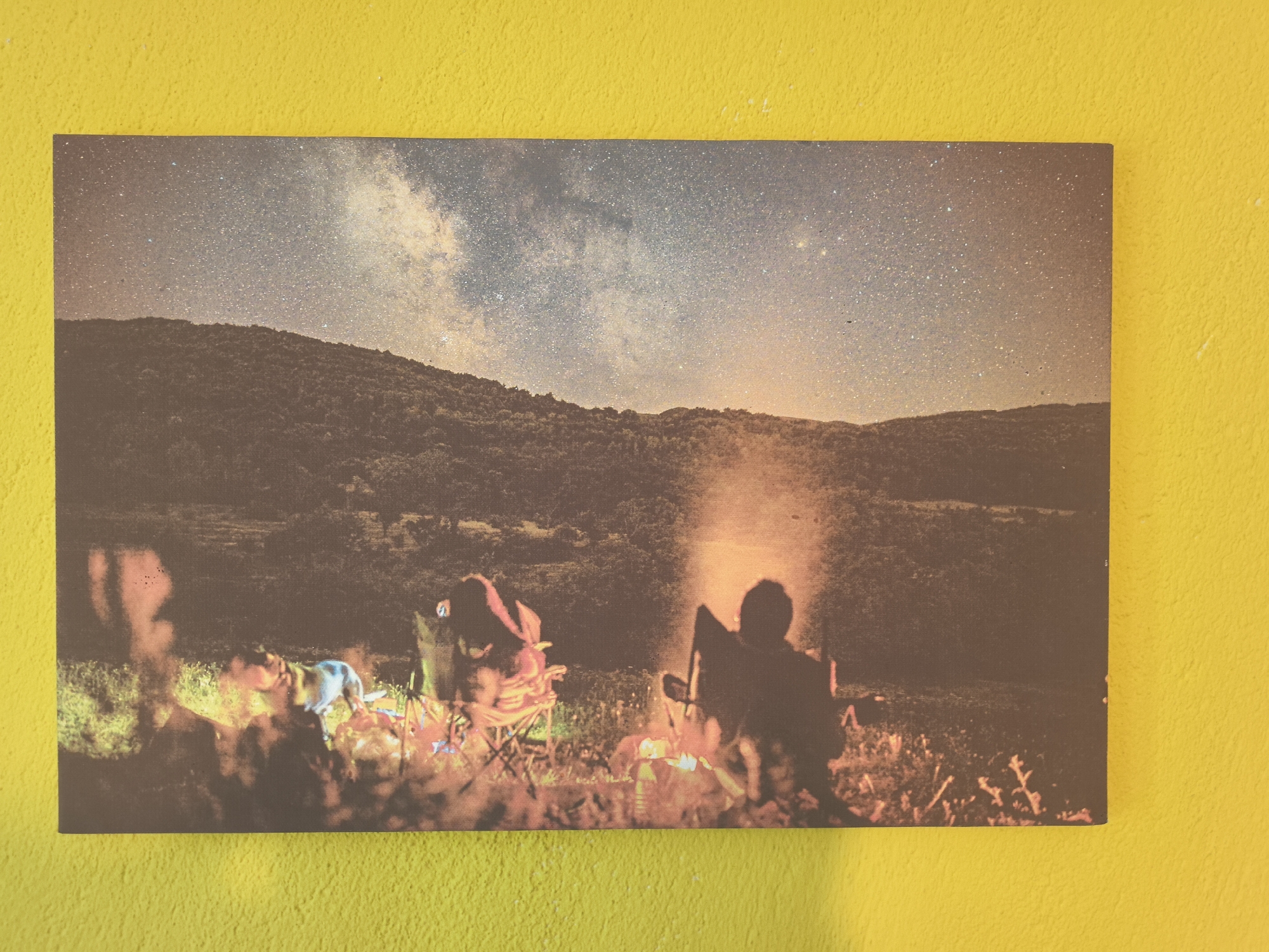 A landscape photograph depicts a nighttime scene of a campsite near Hrabrino, Bulgaria, as evidenced by its distinctive terrain and vegetation. The photo prominently features a silhouette of two figures seated in camping chairs beside a campfire, their faces obscured by the darkness and the flickering flames. A dog is also partially visible near the campfire. The campsite is situated on a grassy field with scattered plants. Above the figures and landscape, the night sky is visible, showcasing a vast expanse of stars, including the Milky Way. The backdrop is composed of a densely forested hill. The photograph appears to be affixed to a yellow wall.