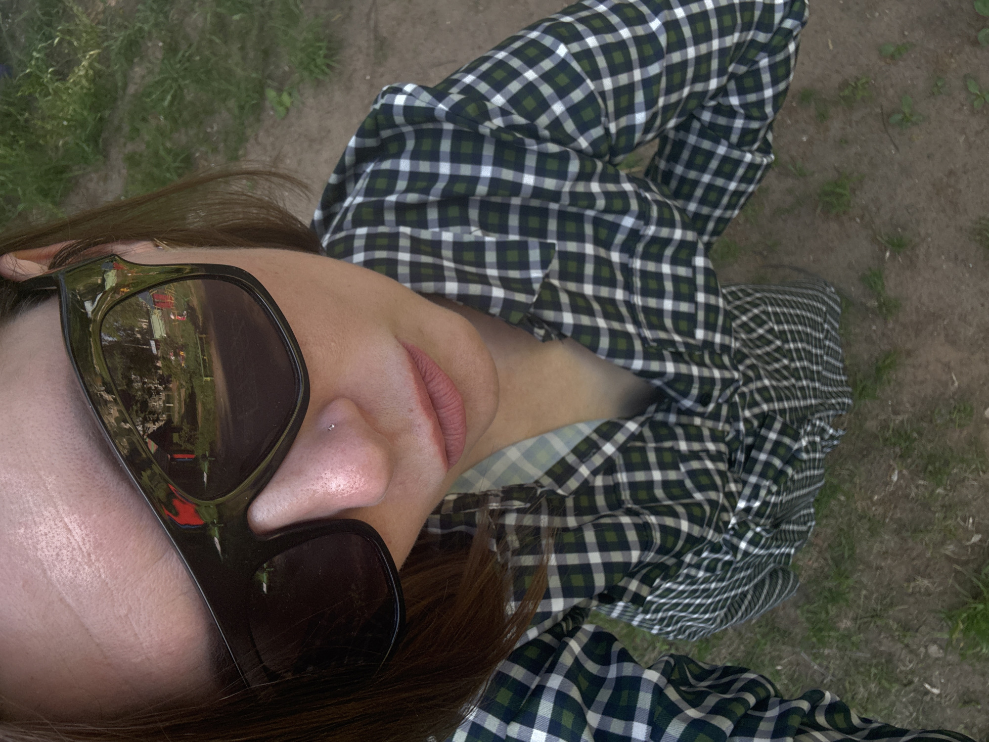 A woman lies on the ground in an outdoor setting in Chaikovsky, Russia, as evident in the sunglasses’ reflection. She wears a green, white, and black checkered long-sleeved shirt. She has brown hair and is wearing sunglasses. She has a piercing on her nose. The ground is a mix of dirt and grass.